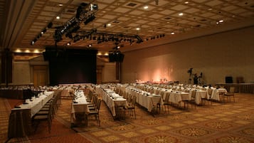 Een ruime congreszaal met rijen tafels en stoelen, een podium met lichtinstallatie en een kamerbreed tapijt met patroon.