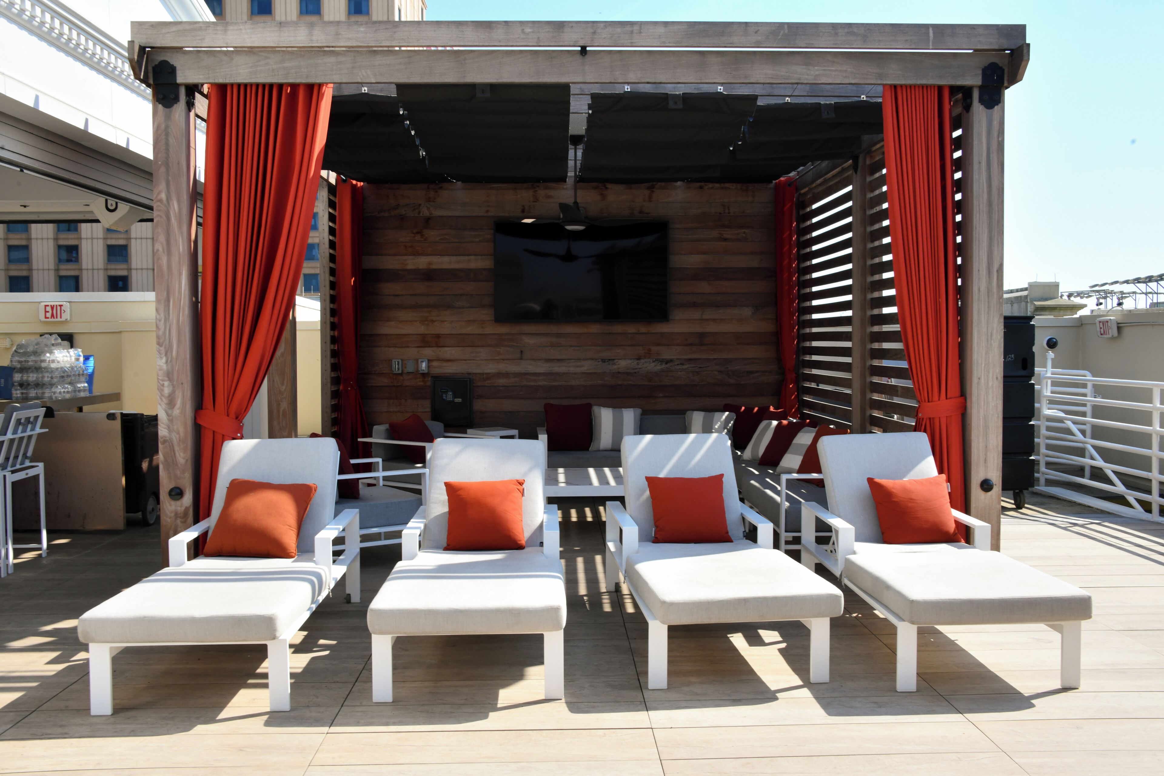 A rooftop lounge area with white lounge chairs and red cushions, a wooden wall with a mounted TV, and a clear sky.