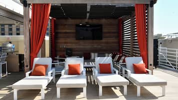 A rooftop lounge area with white lounge chairs and red cushions, a wooden wall with a mounted TV, and a clear sky.