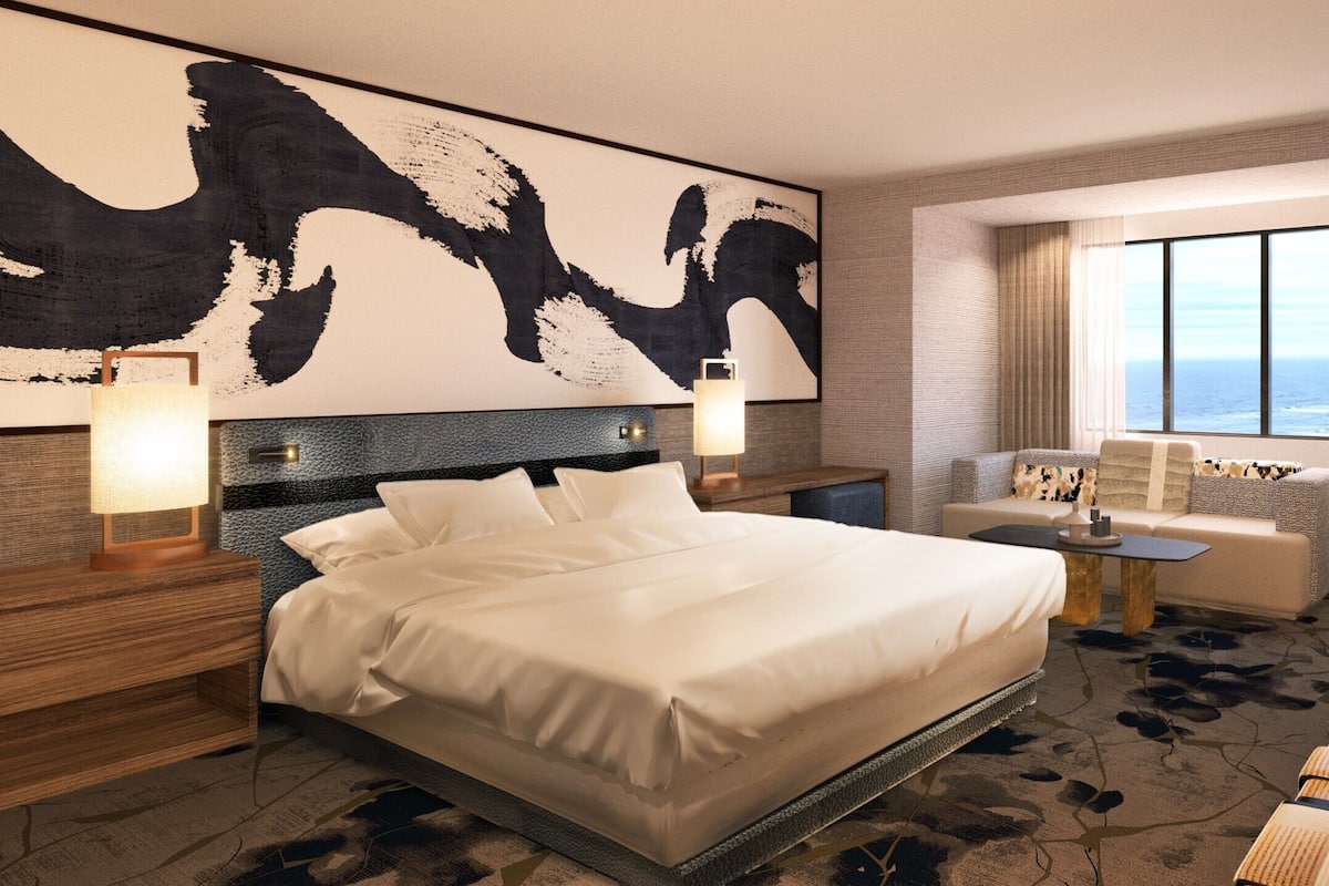 Ein Hotelzimmer mit einem großen Bett, einer Sitzecke mit Meerblick und einem großen, abstrakten Gemälde an der Wand.