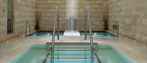 A large indoor swimming pool with a central waterfall feature, surrounded by stone walls and equipped with metal ladders.