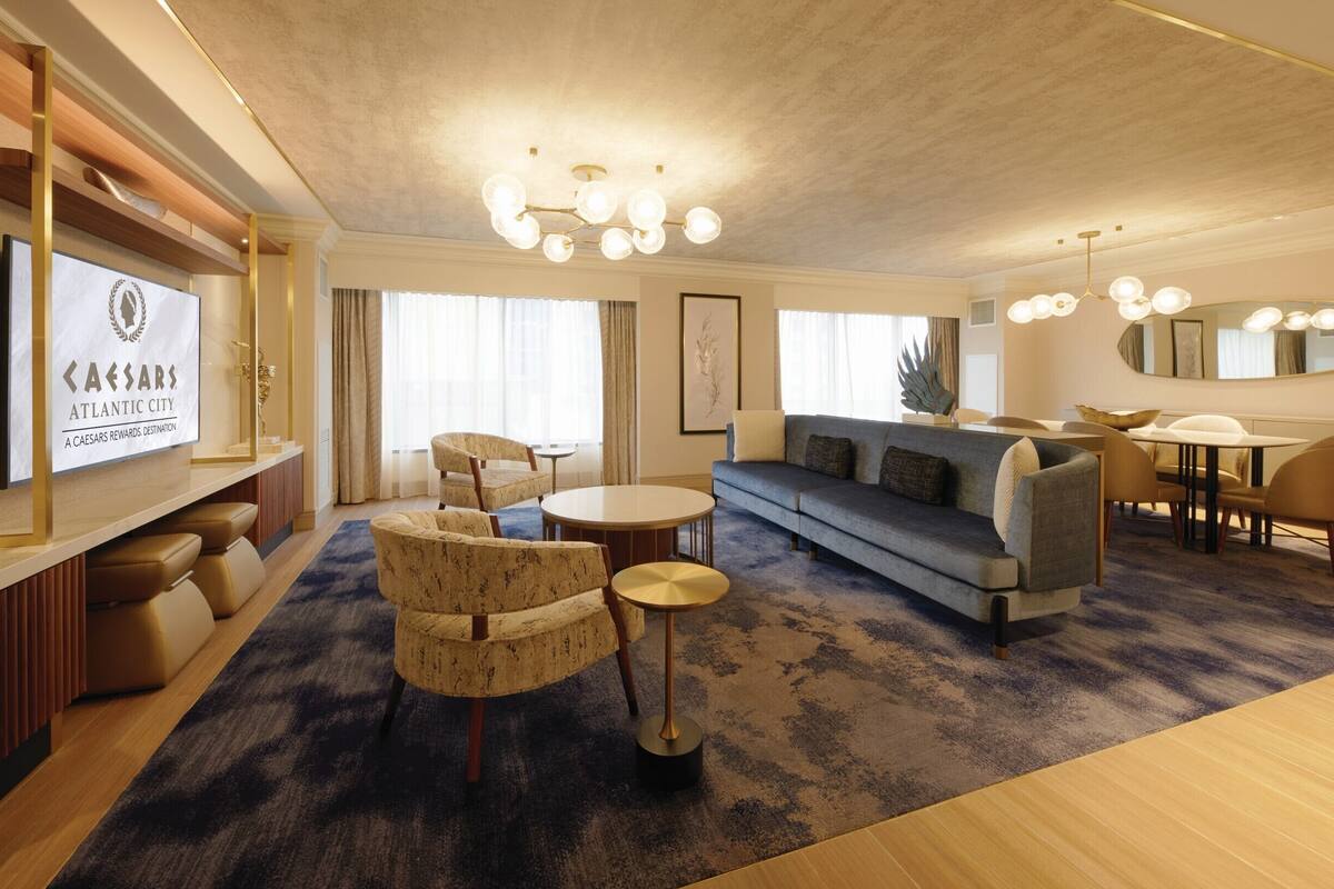 Ein modernes Hotelzimmer mit einer Couch, Sesseln, einem Esstisch und einem Fernseher, der ein Schild mit dem Caesars Rewards Destinations-Logo zeigt.