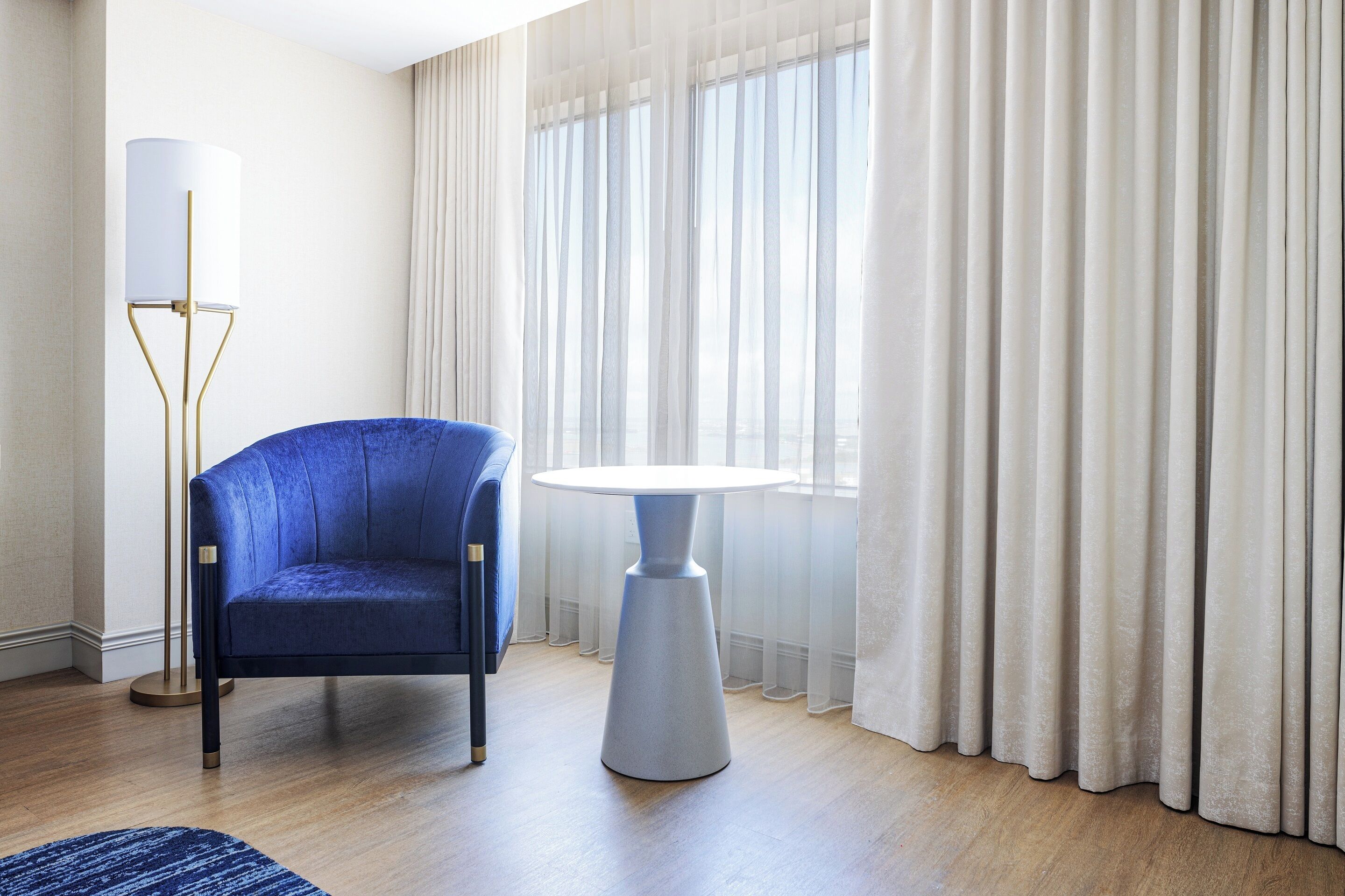 A room with a blue armchair, a round table, a floor lamp, and sheer curtains.