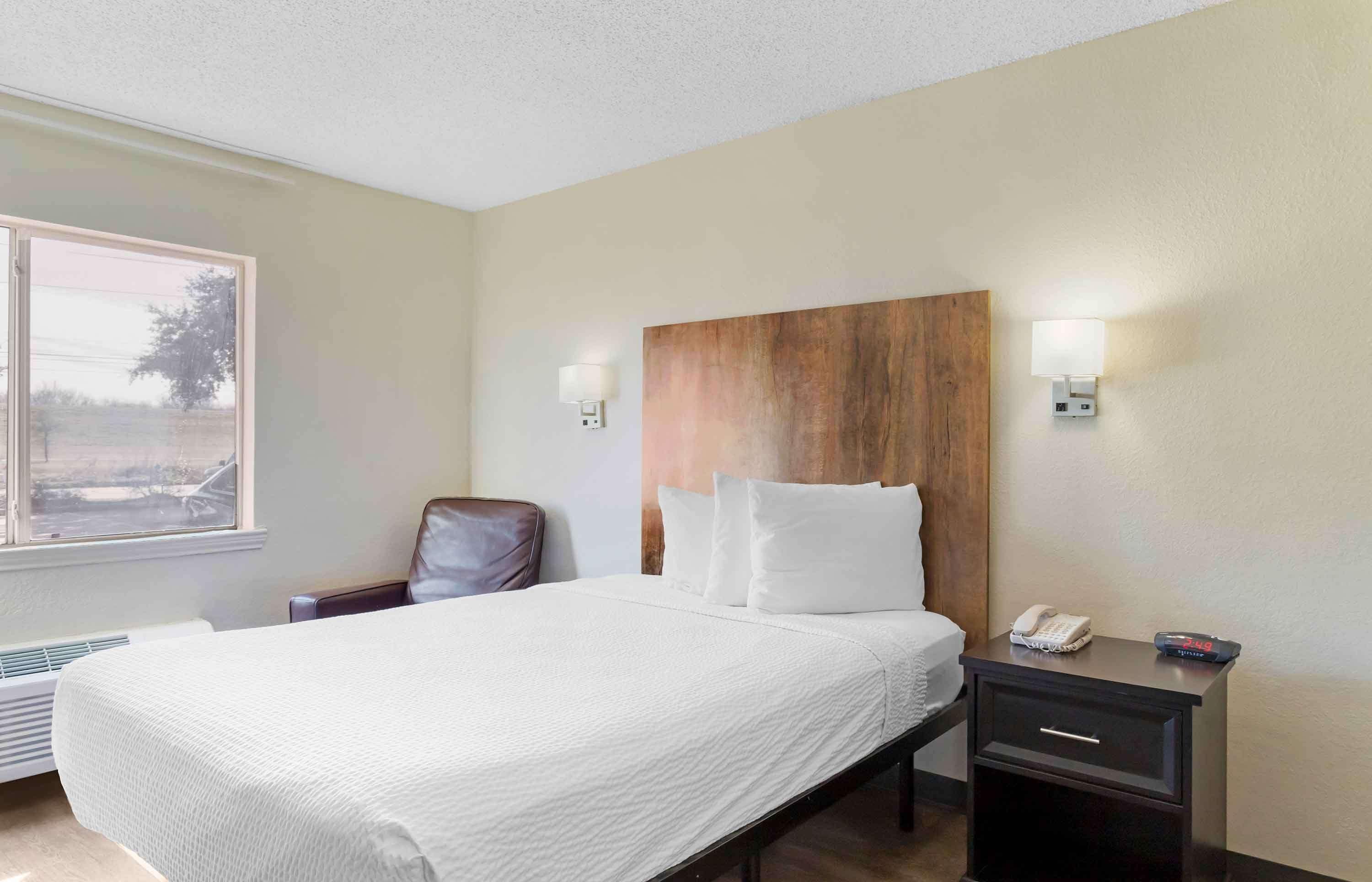 A hotel room with a large bed, a wooden headboard, two wall-mounted lamps, a bedside table with a phone and remote control, and a chair.