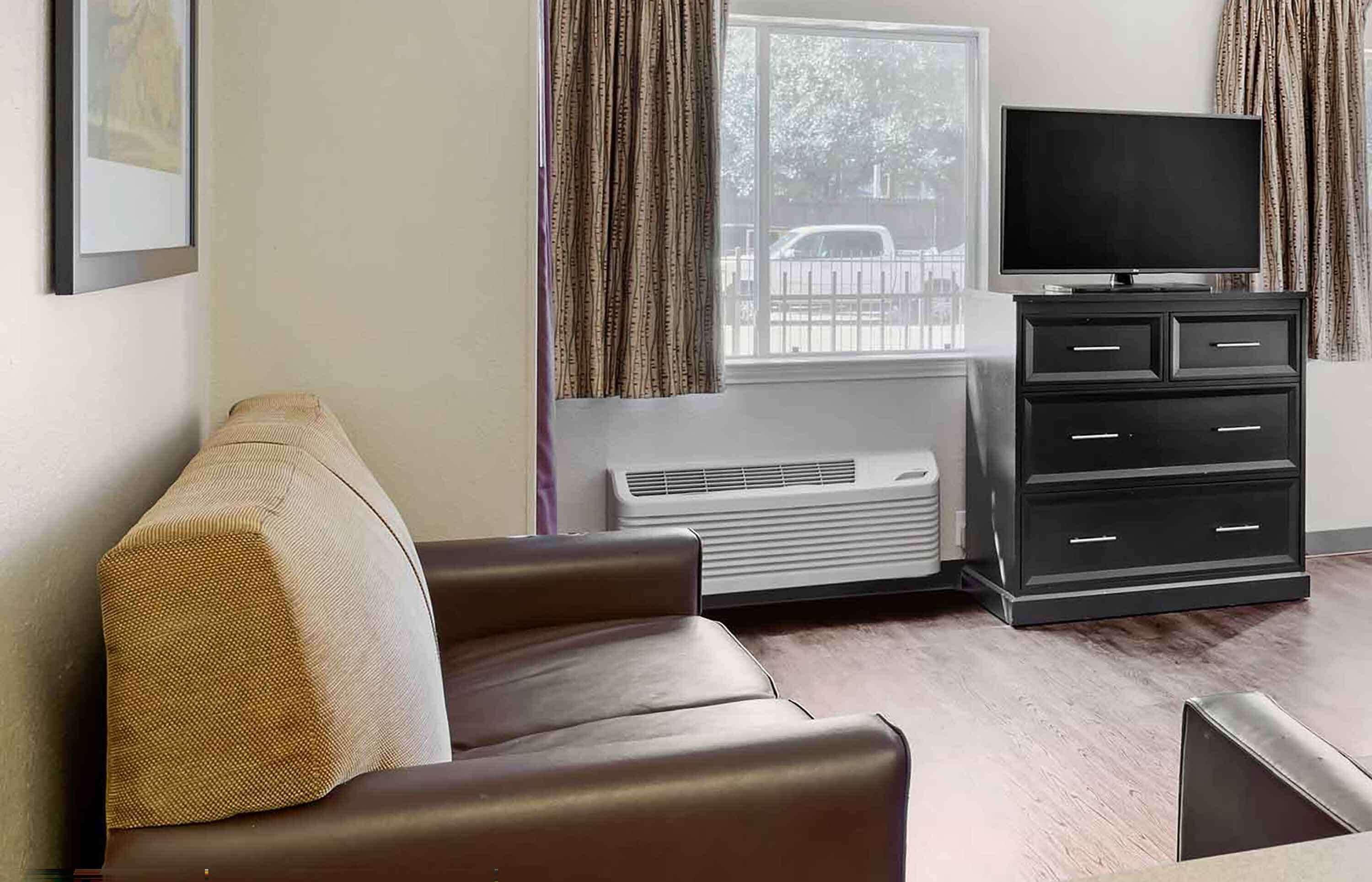 A living room with a brown leather sofa, a black entertainment unit with a flat-screen TV, and a window with curtains.