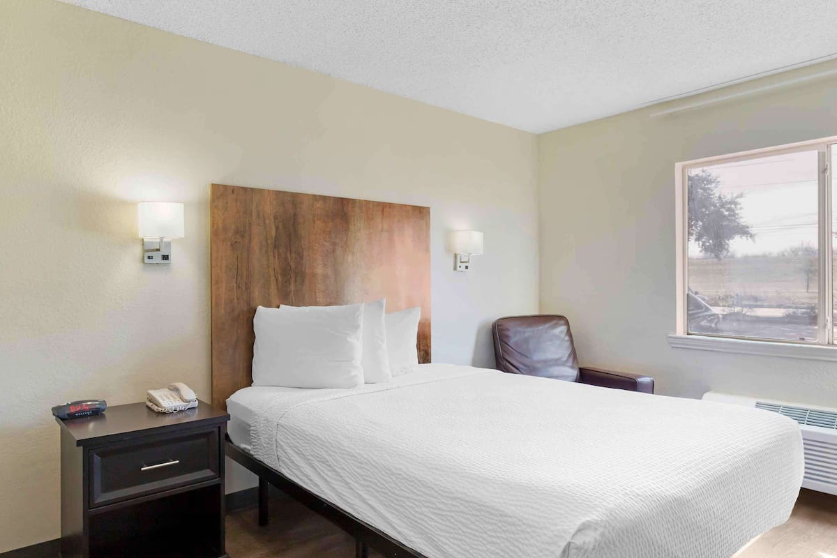 A hotel room with a large bed, a wooden headboard, bedside lamps, a chair, a nightstand with a phone, and a window with a view.