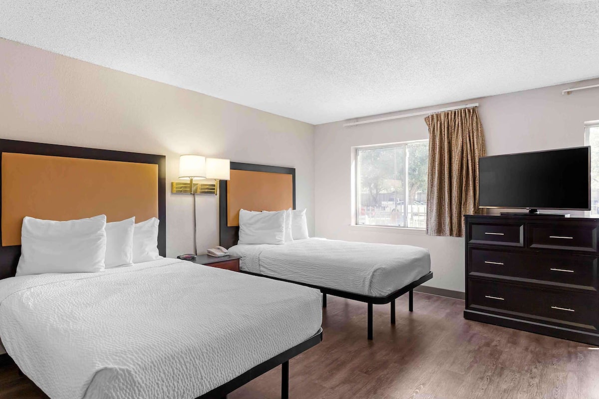 A hotel room with two beds, a TV, a dresser, and a window with curtains.