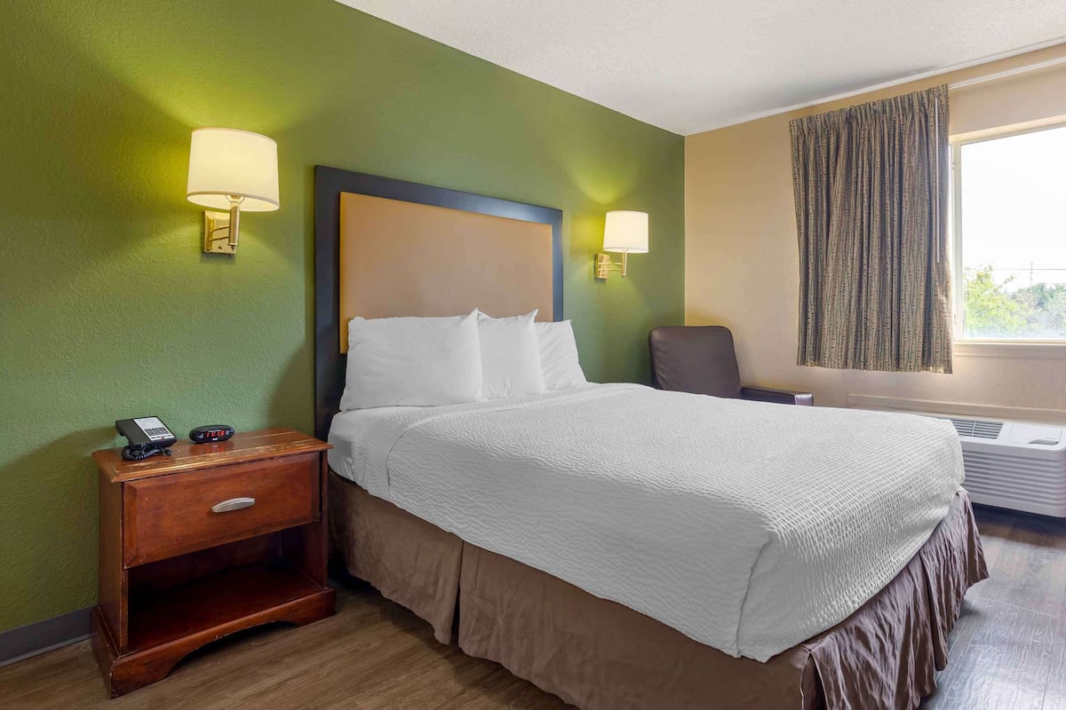 A hotel room with a bed, bedside lamps, a nightstand, a chair, and an air conditioning unit.