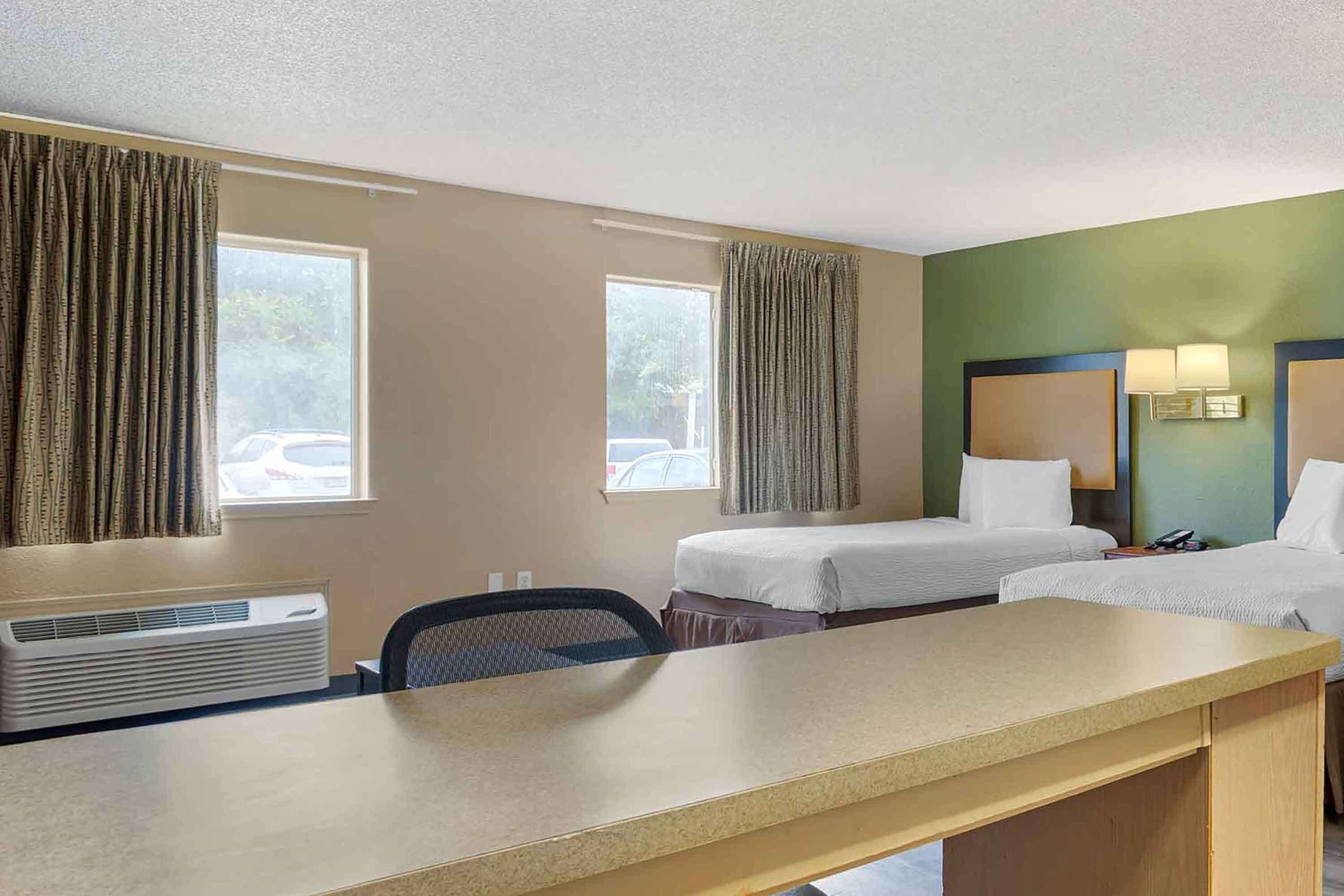 A hotel room with two single beds, a desk, a chair, an air conditioning unit, and a view of the outside.