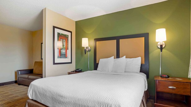 A hotel room with a large bed, two bedside tables, a nightstand, a chair, and a framed picture on the wall. - Extended Stay America Select Suites - Houston - Sugar Land (Sugar Land)