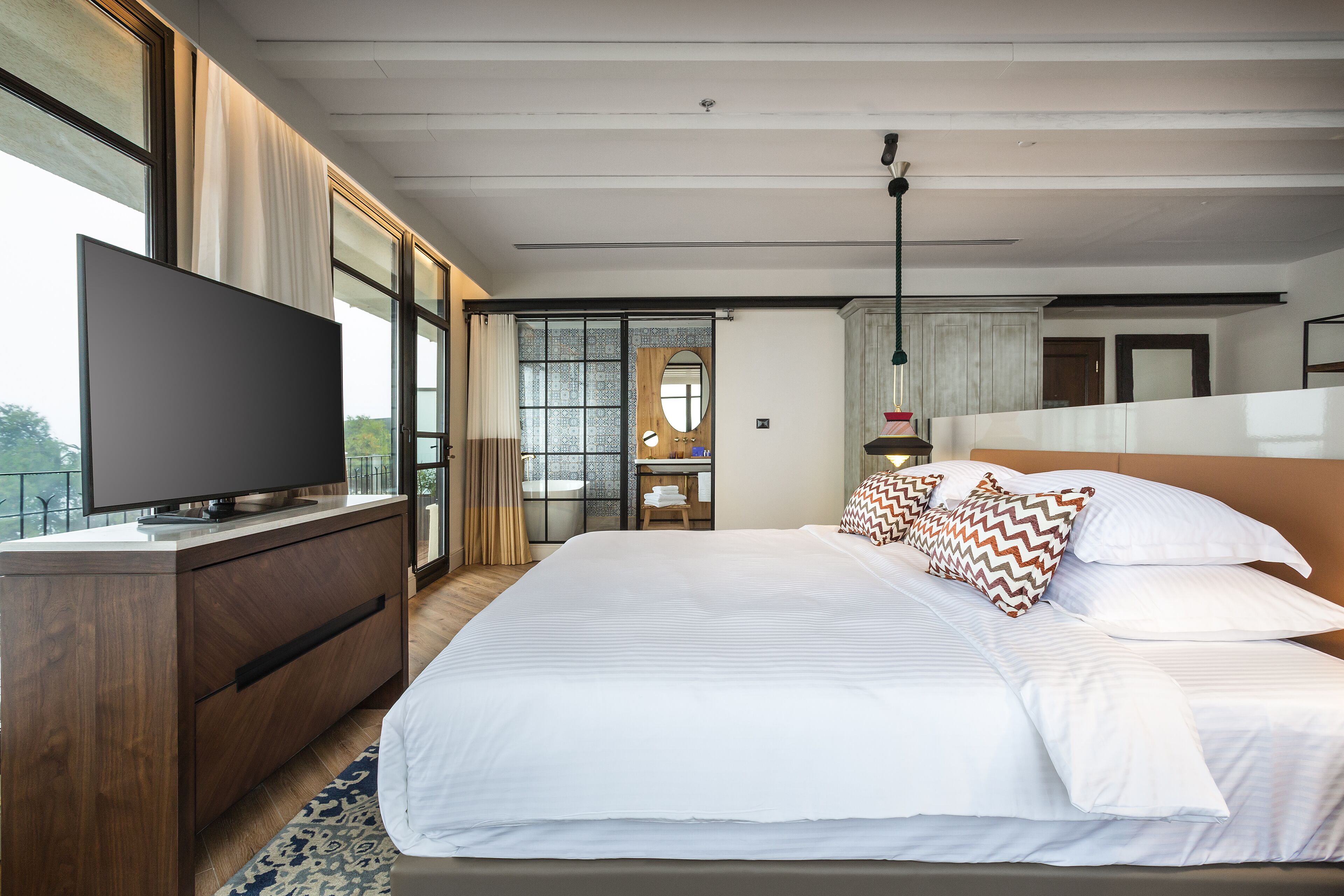 A modern hotel room with a large bed, a flat-screen TV, a wooden dresser, and a view of the outdoors through large windows.
