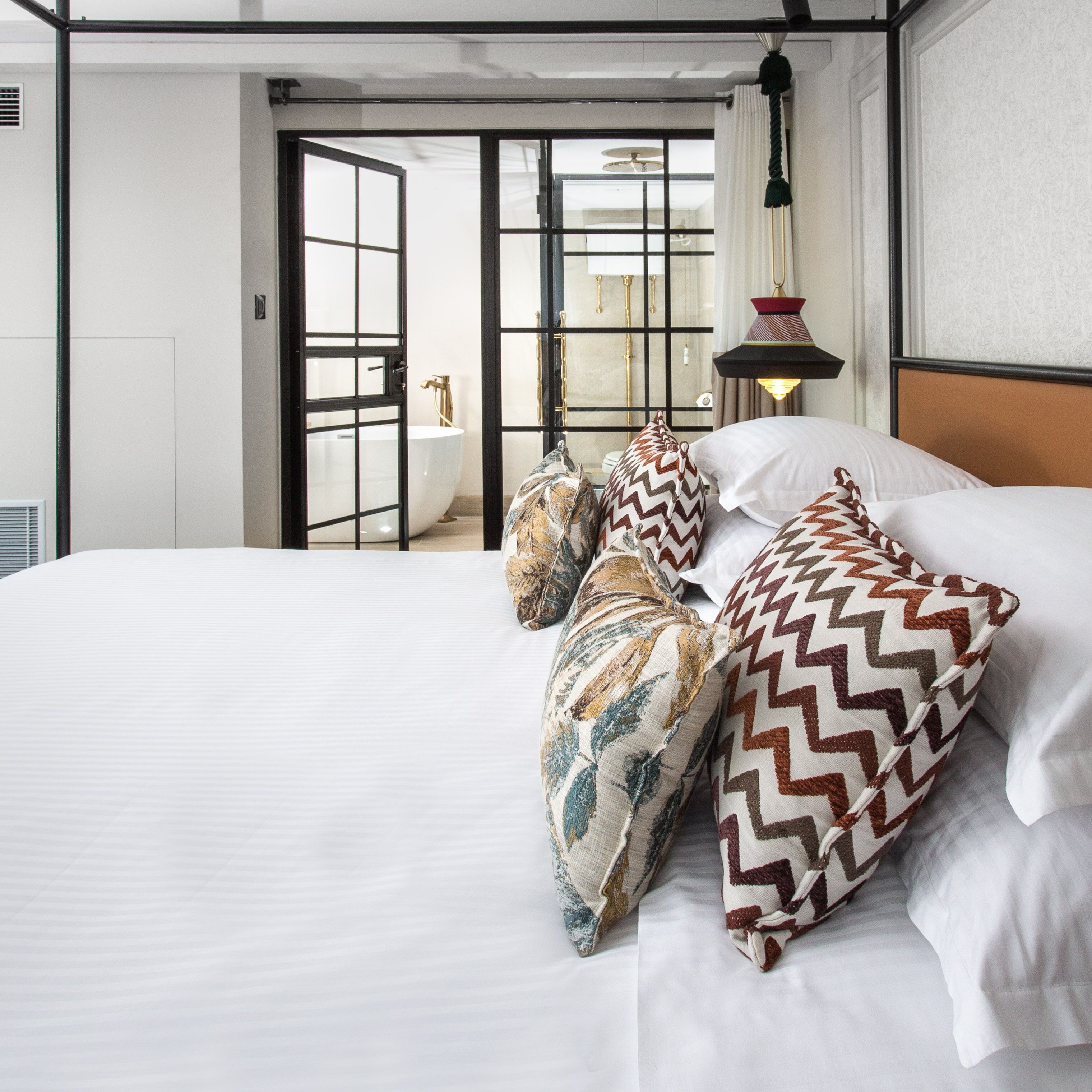 Une chambre d’hôtel avec un grand lit, des oreillers à motifs, une lampe de chevet et une vue sur la salle de bain par une porte ouverte.