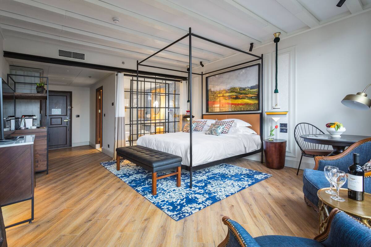A modern bedroom with a four-poster bed, a dining area with a table and chairs, and a kitchen in the background.