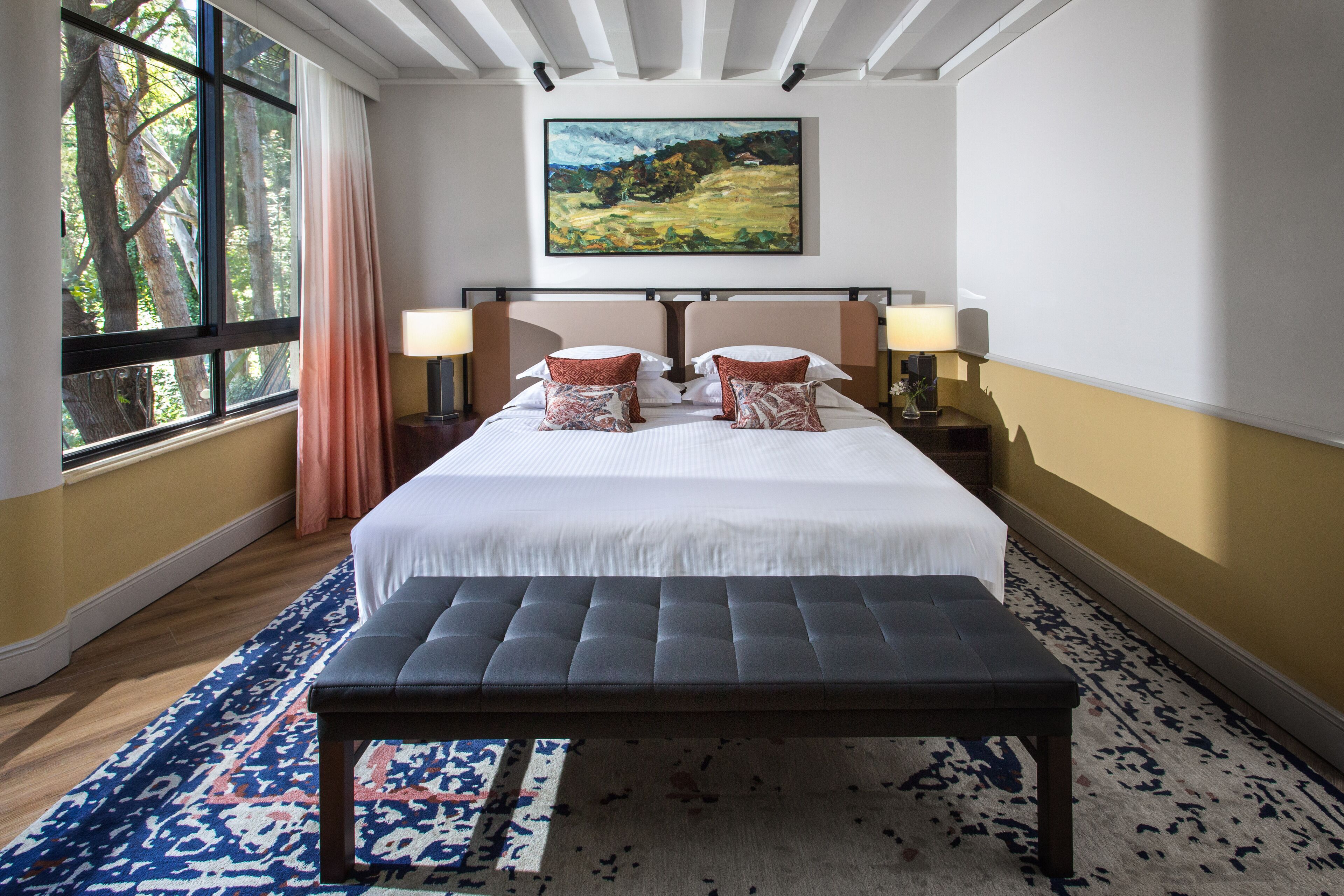 A hotel room with a large bed, two bedside tables with lamps, a bench, a large window with curtains, and a painting on the wall.