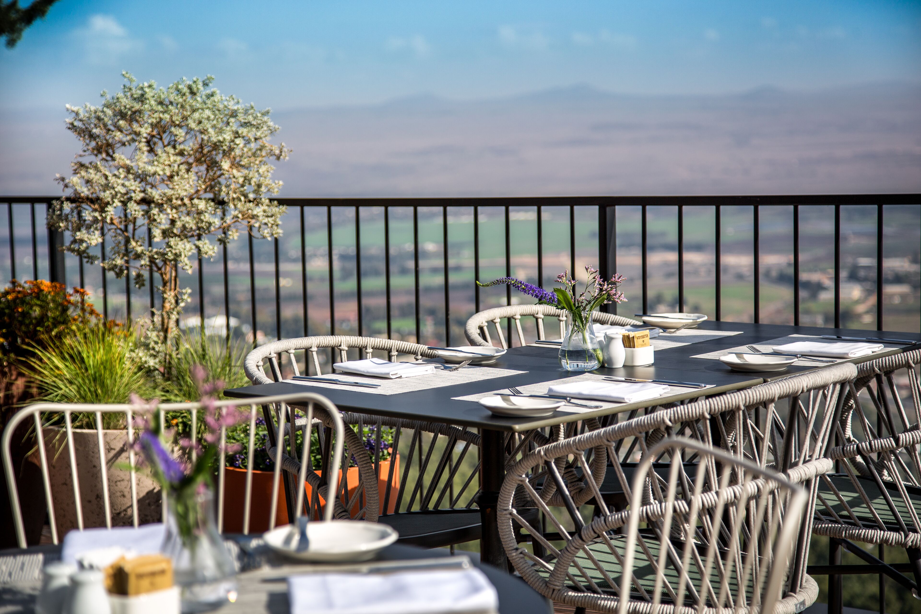 Une table à manger installée à l’extérieur, avec des chaises en osier, un vase rempli de fleurs et une vue sur la campagne.