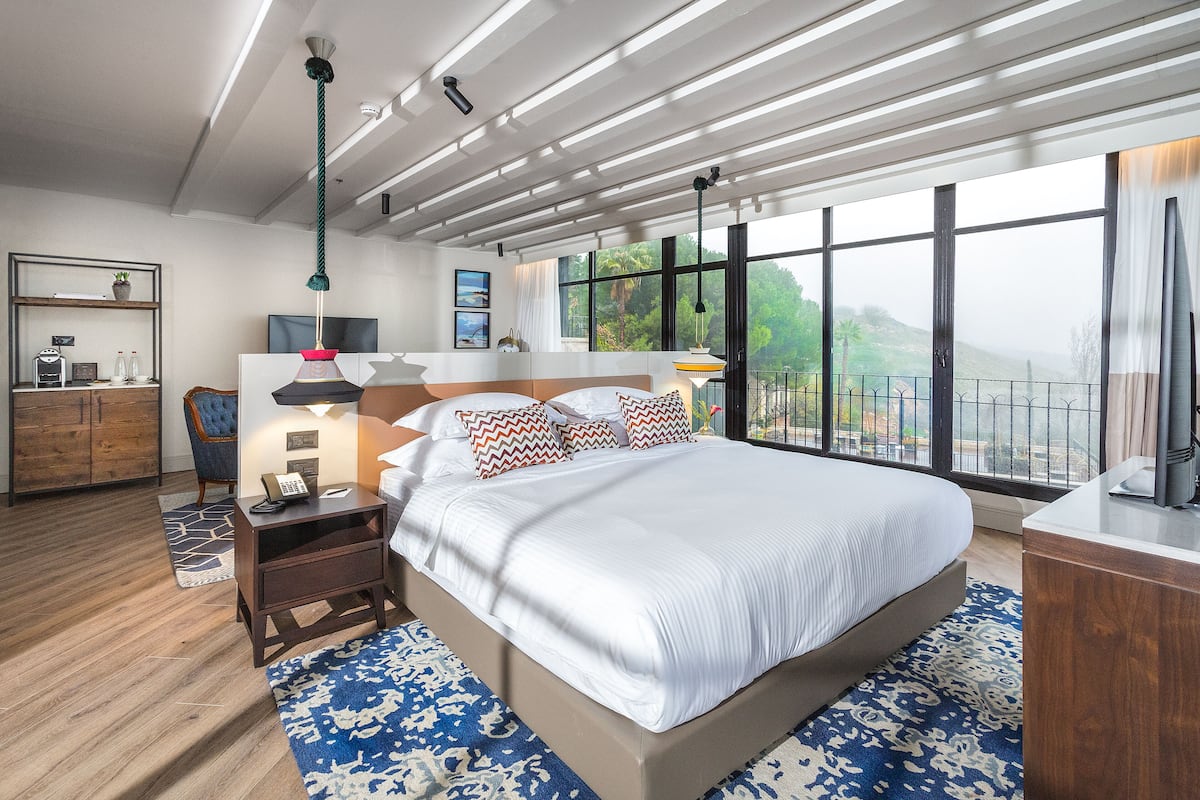 A modern hotel room with a large bed, a desk, a chair, a TV, and a large window with a view of the outdoors.