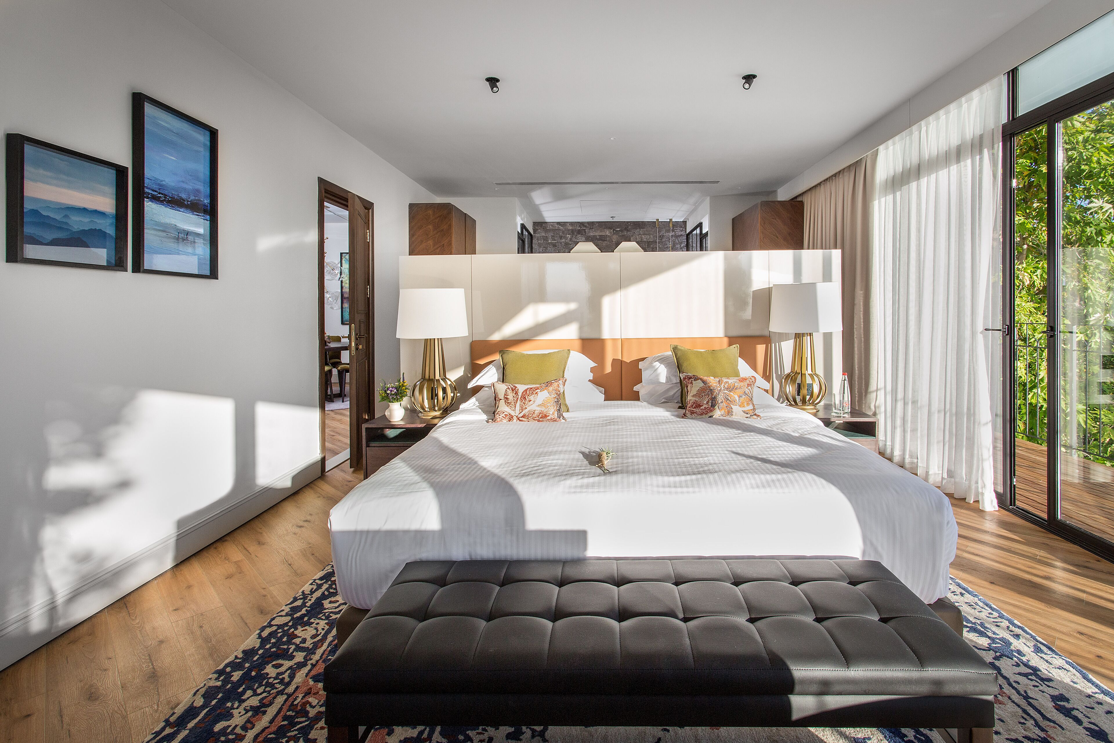 A spacious bedroom with a large bed, a bench, and a view of greenery through the window.