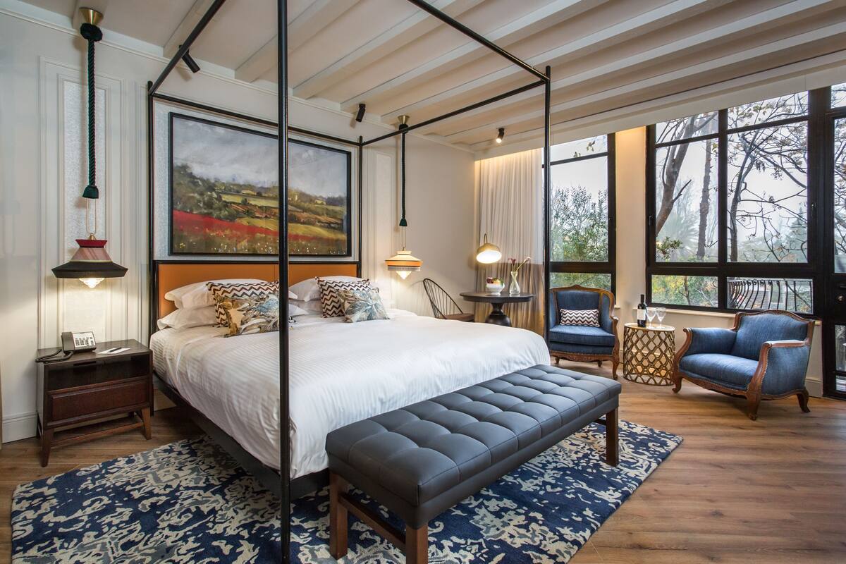 A bedroom with a four-poster bed, a bench, a chair, and a nightstand with a lamp.
