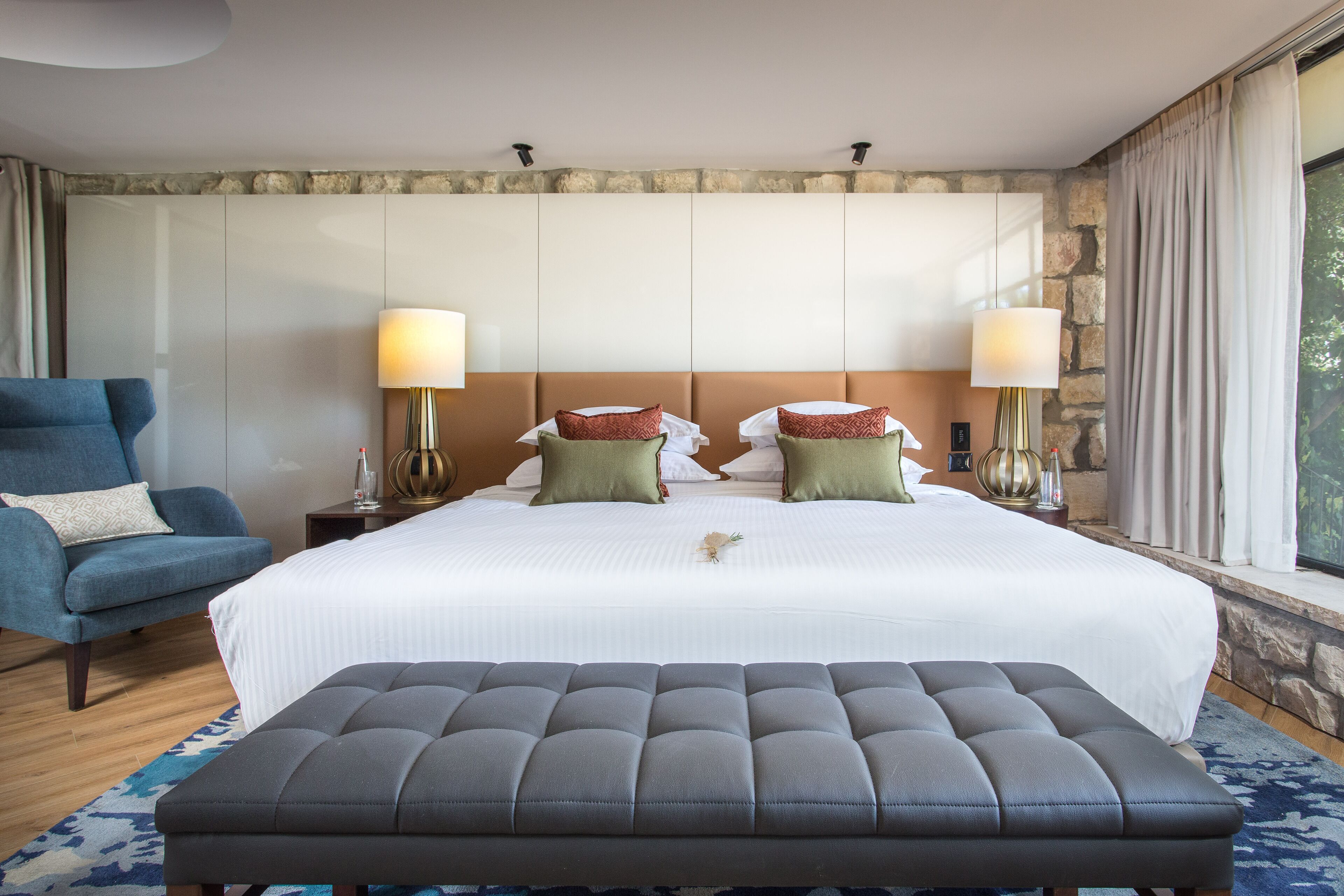 Ein Hotelzimmer mit einem großen Bett, einem blauen Sessel, einer Bank und einer Wand mit Steinverkleidung.