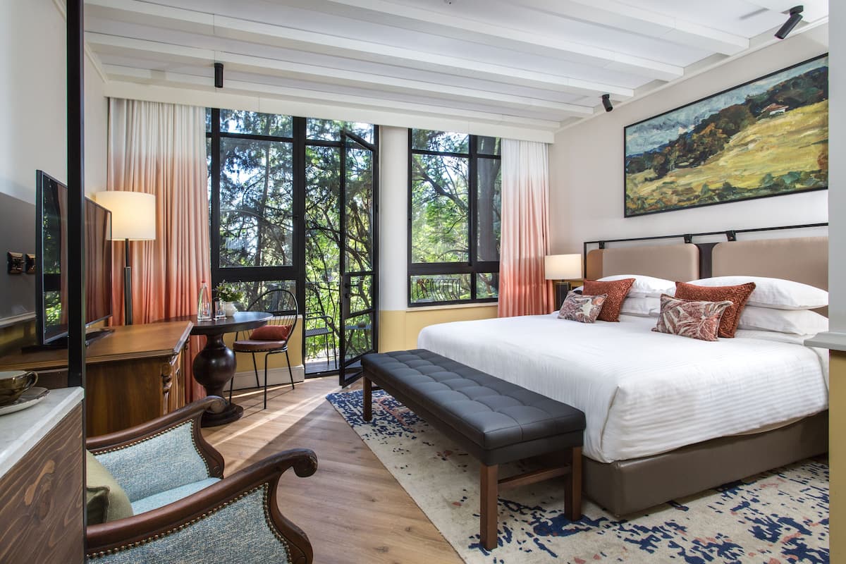 A bedroom with a large bed, a desk, a chair, and a bench. There are large windows with curtains, a painting on the wall, and a view of trees outside.