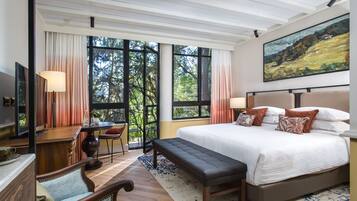 A bedroom with a large bed, a desk, a chair, and a bench. There are large windows with curtains, a painting on the wall, and a view of trees outside.