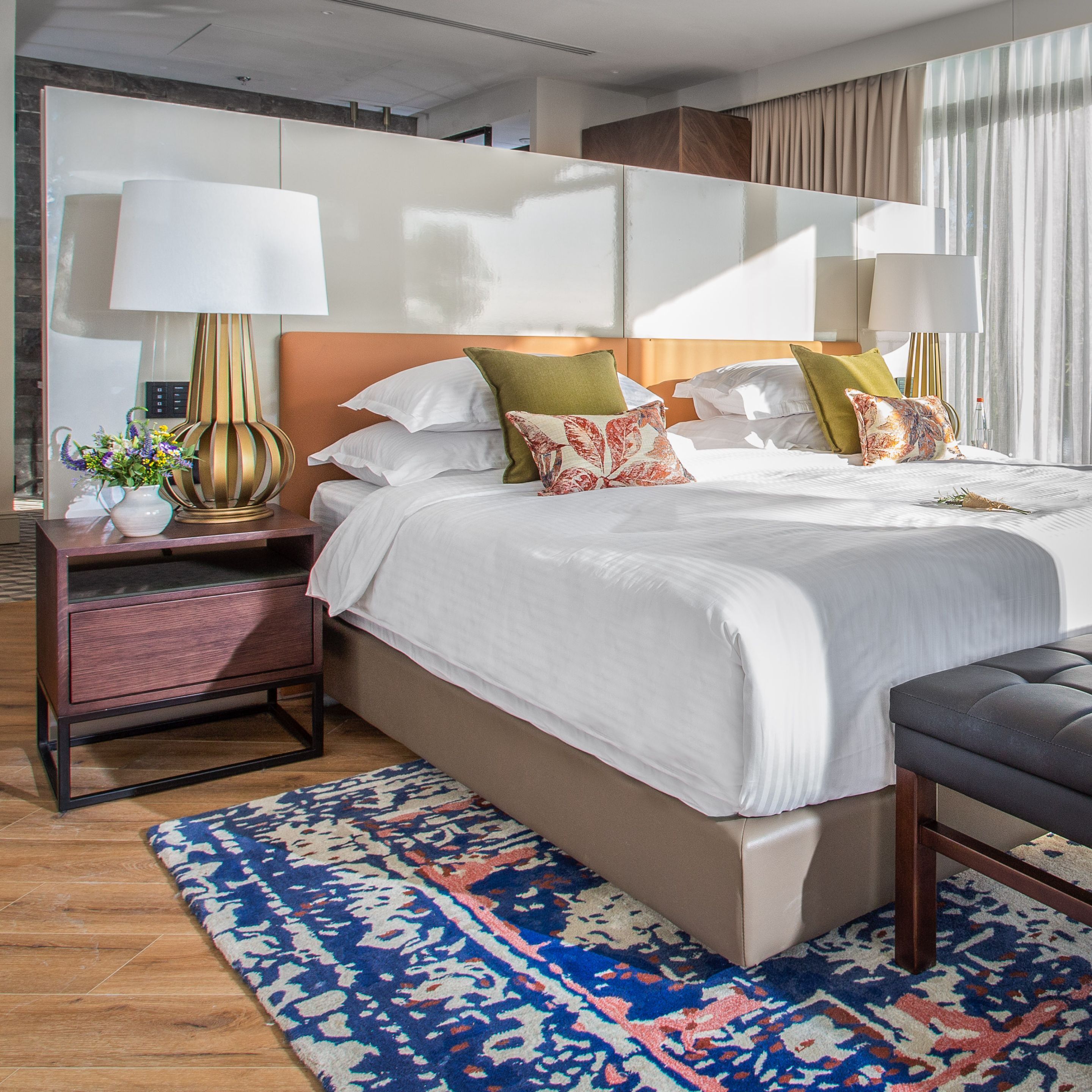 A modern hotel room with a large bed, two bedside tables with lamps, a small side table with a vase, and a patterned rug.