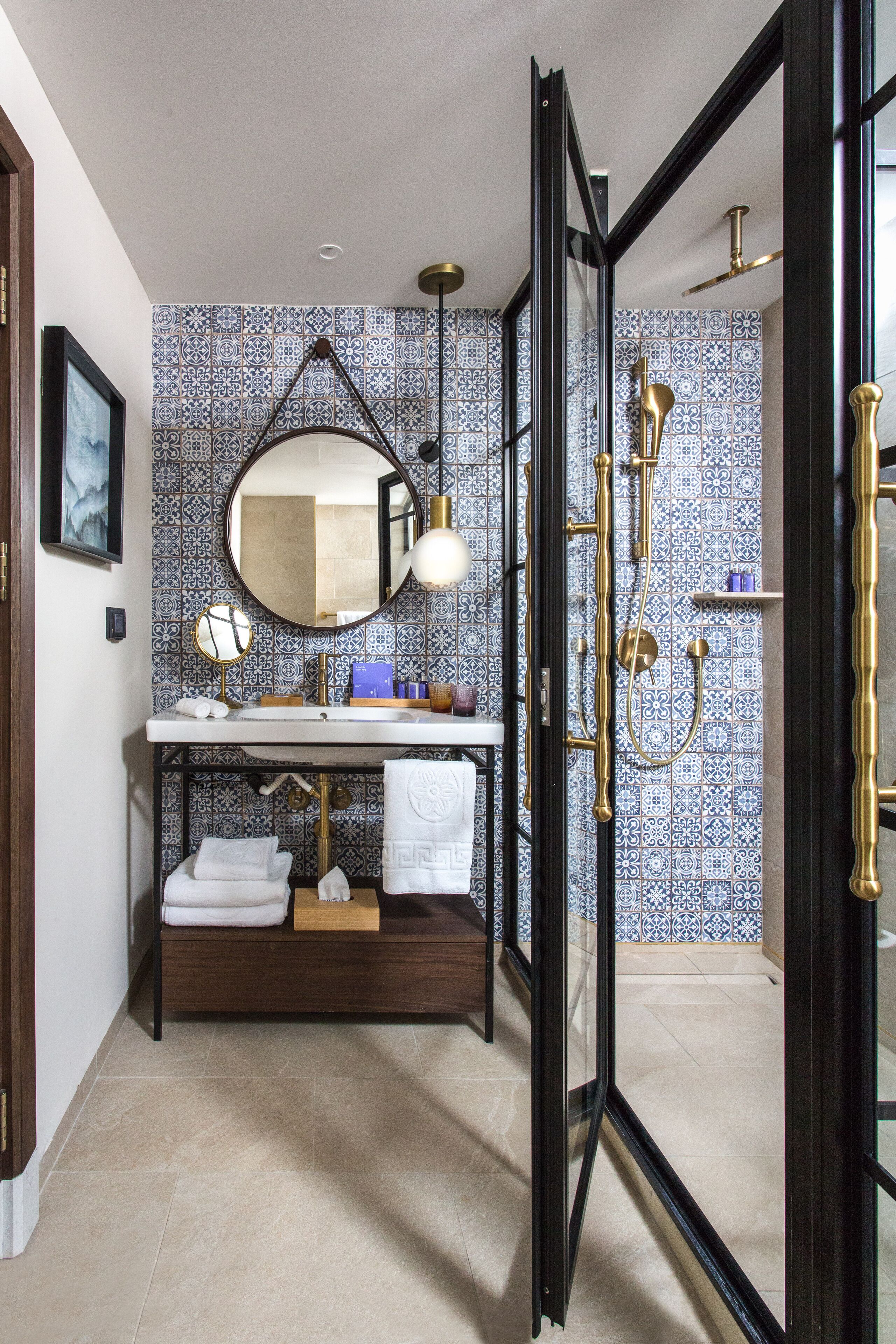 A bathroom with a tiled backsplash, a round mirror, a sink with a towel rack, and a shower area.