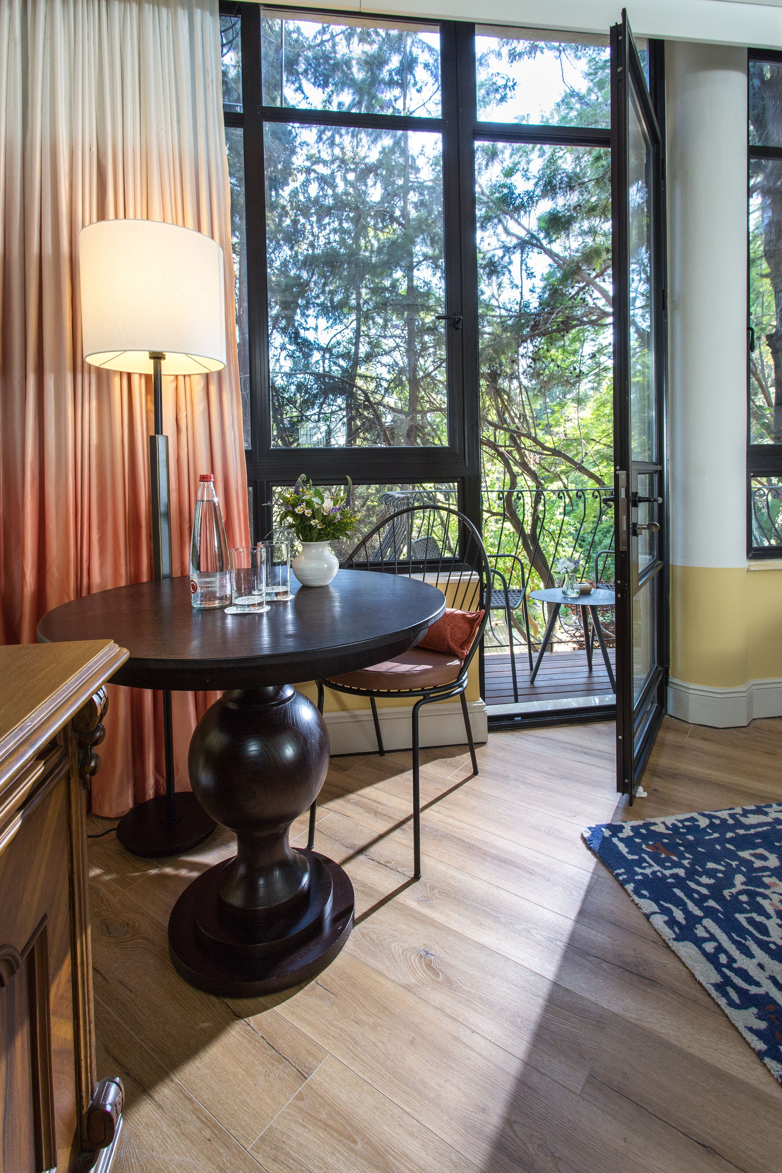 A dining area with a table, chairs, and a lamp. There is a view of trees through large windows.