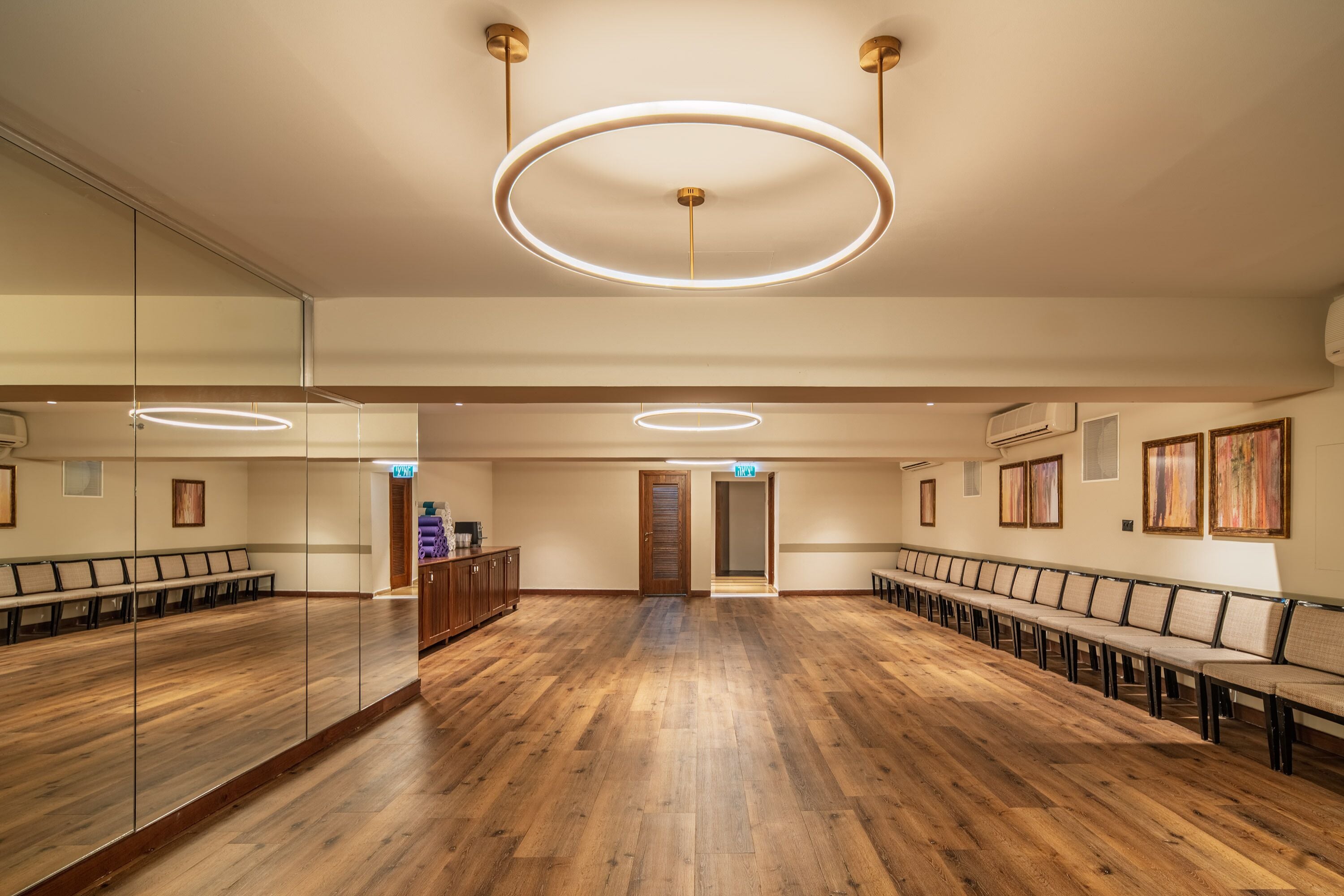 A spacious room with wooden flooring, a long row of chairs, and a large mirror on one wall.