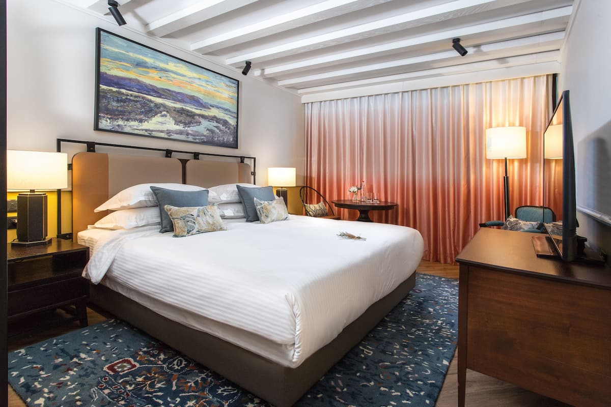 A hotel room with a large bed, two bedside tables with lamps, a desk, a chair, a television, and a painting on the wall.