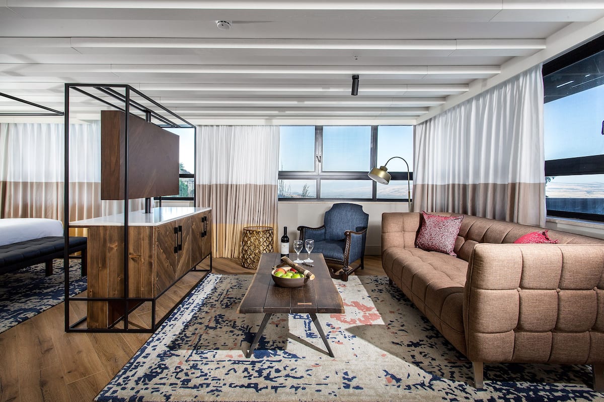 A modern hotel room with a wooden desk, a sofa, a dining table with a bowl of fruit, and a large window offering a view.