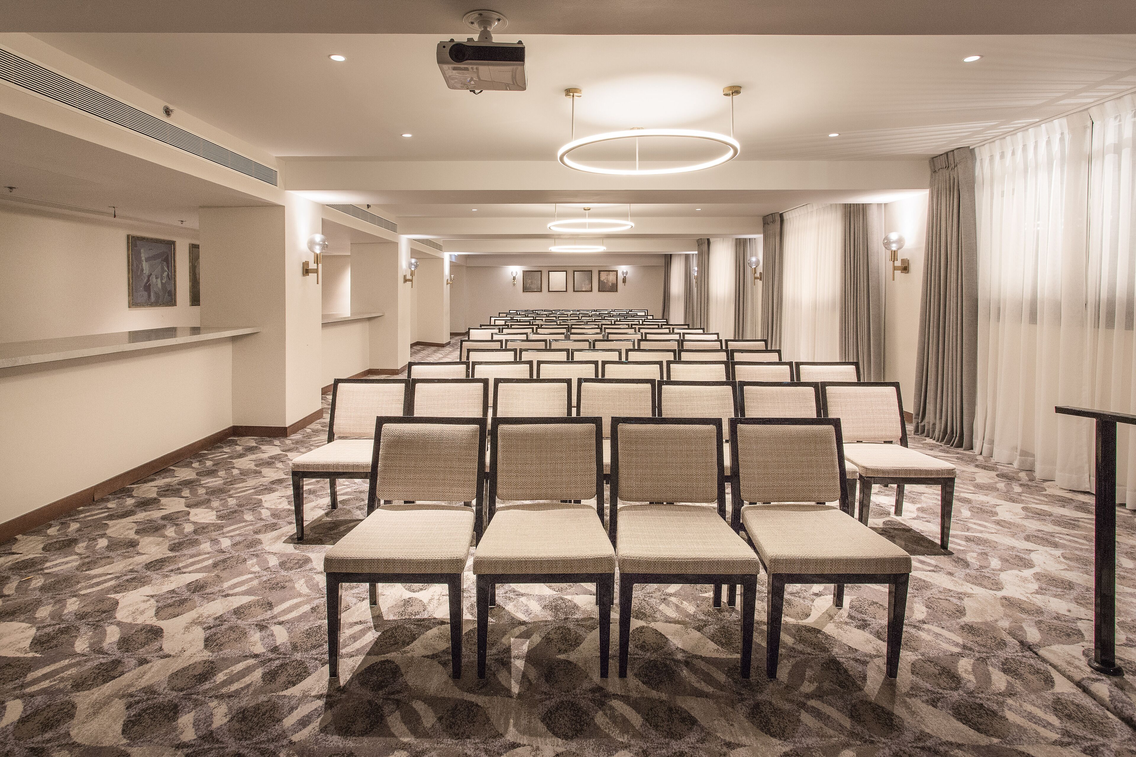 Un salón de conferencias con filas de sillas, piso alfombrado y un proyector en el techo.