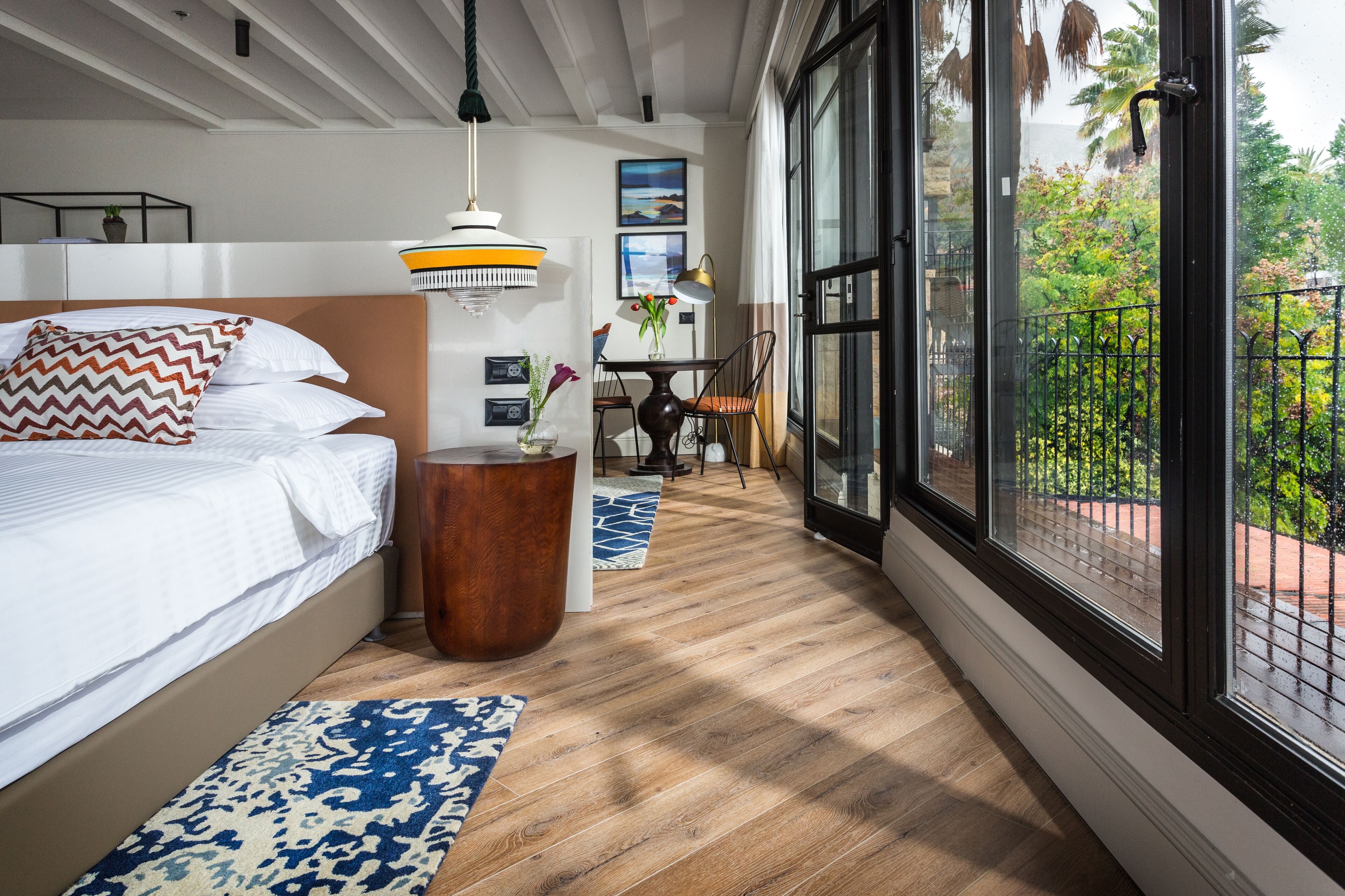 A bedroom with a bed, a wooden side table, a hanging light fixture, and a view of a balcony with plants.