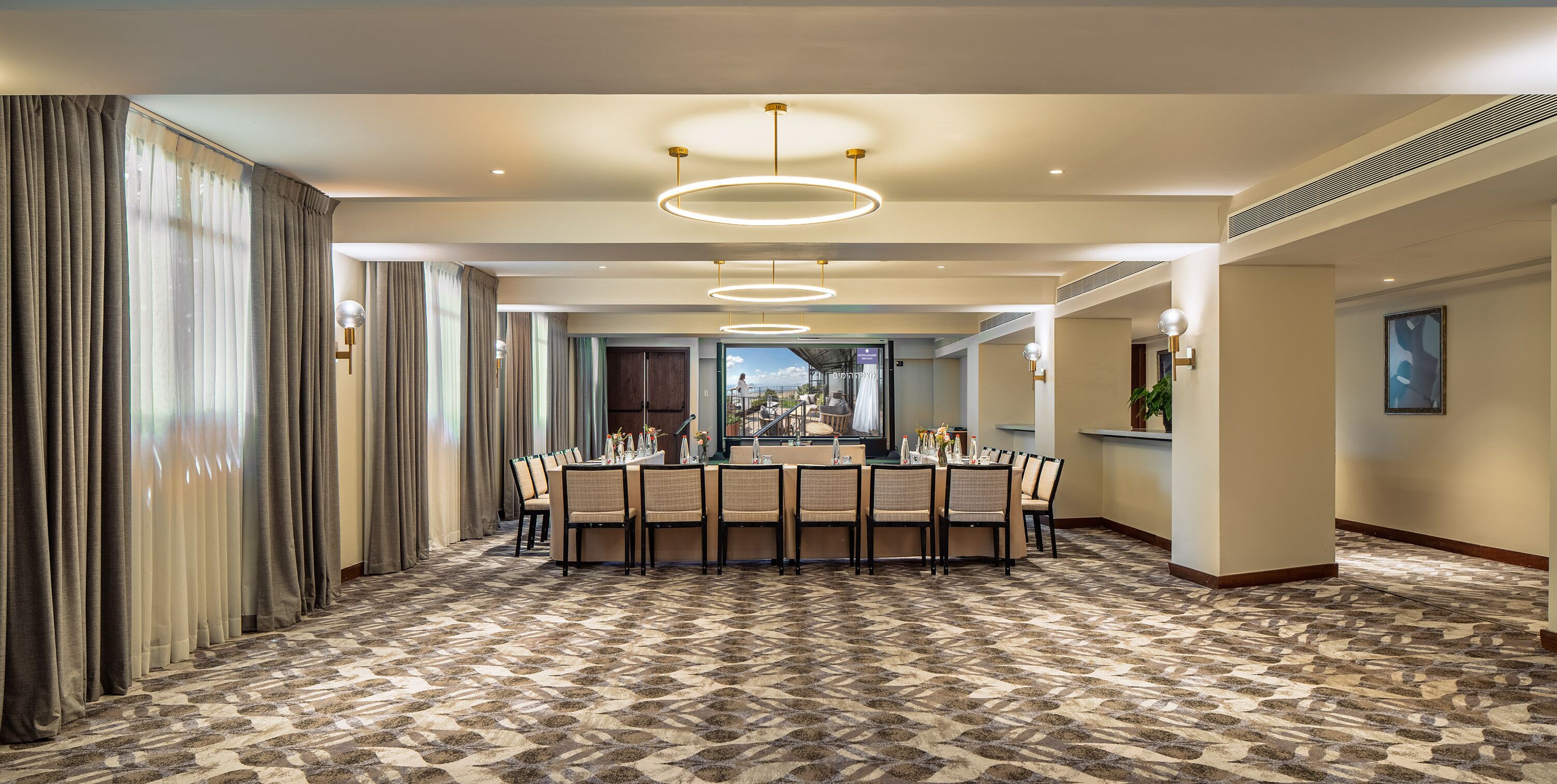 A spacious dining area with a large table set for six, surrounded by chairs, in a hotel with a patterned carpet and modern lighting fixtures.