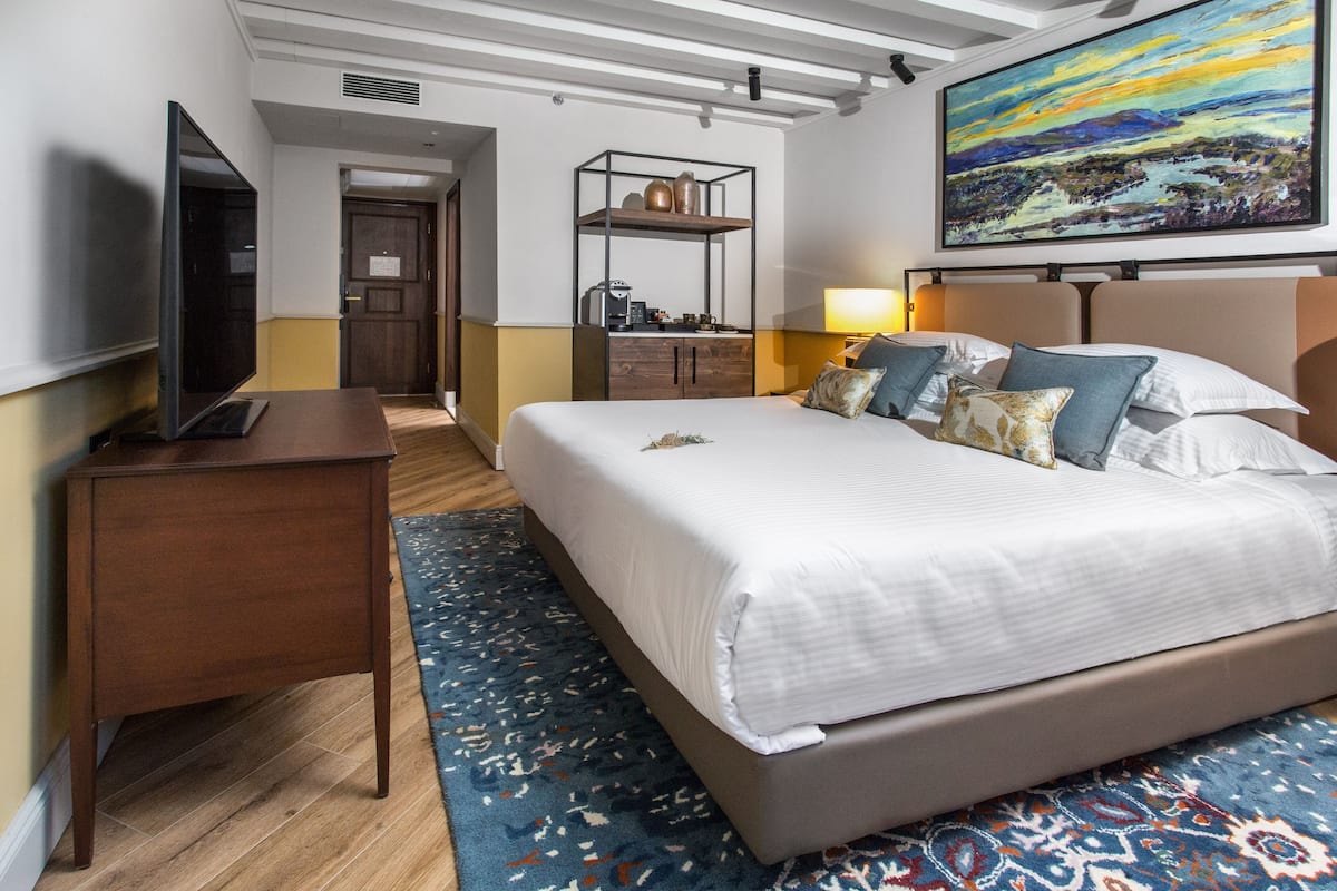 A hotel room with a large bed, a flat-screen TV, a wooden nightstand, a desk, a desk lamp, a bookshelf, and a painting on the wall.