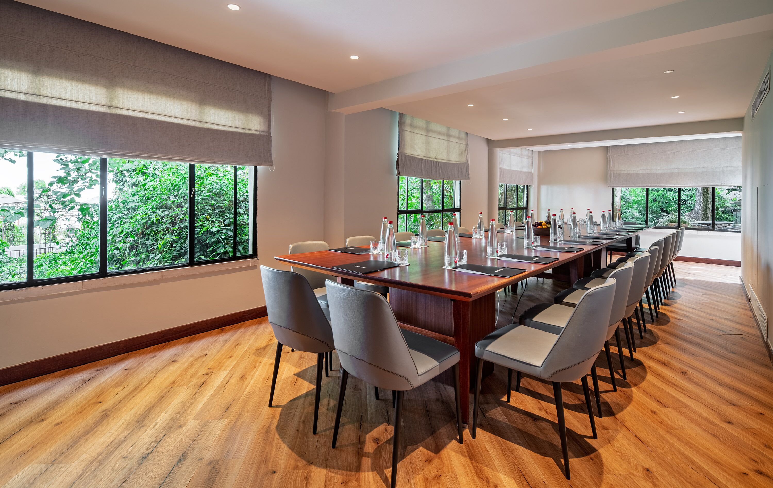 A conference room with a long table, chairs, and large windows offering a view of greenery.