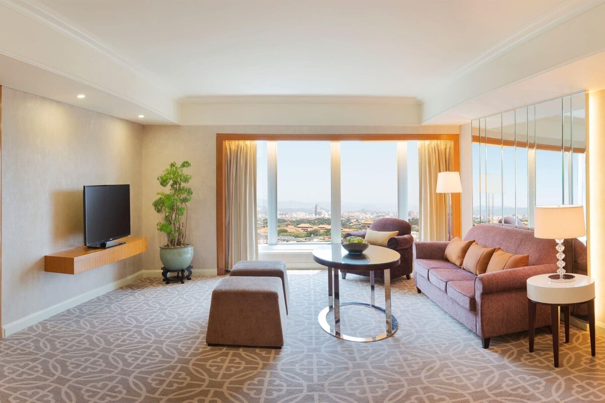A hotel room with a bed, a TV, a sofa, a table, a plant, and a view of the city.