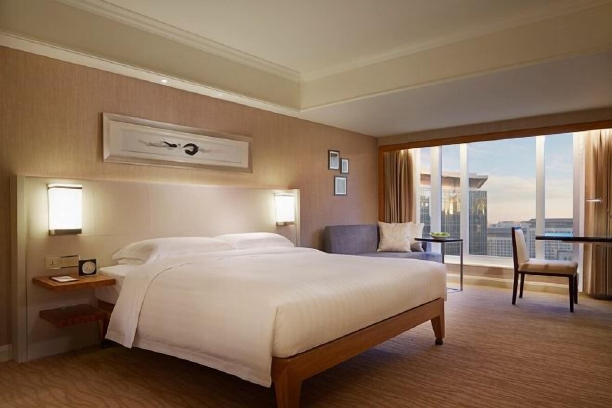 A hotel room with a large bed, a desk with a lamp, a chair, and a view of the city through large windows.
