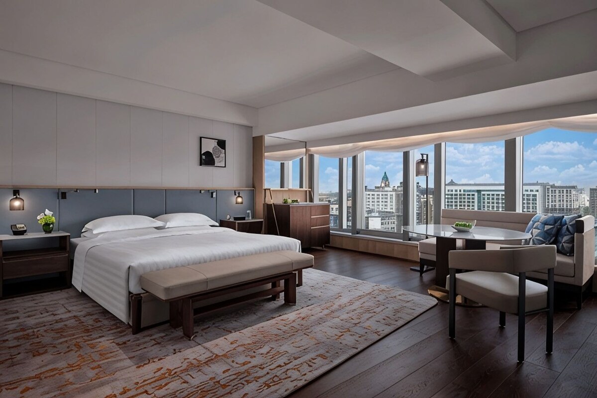 A modern hotel room with a large bed, bedside tables, a desk, and a view of the city.