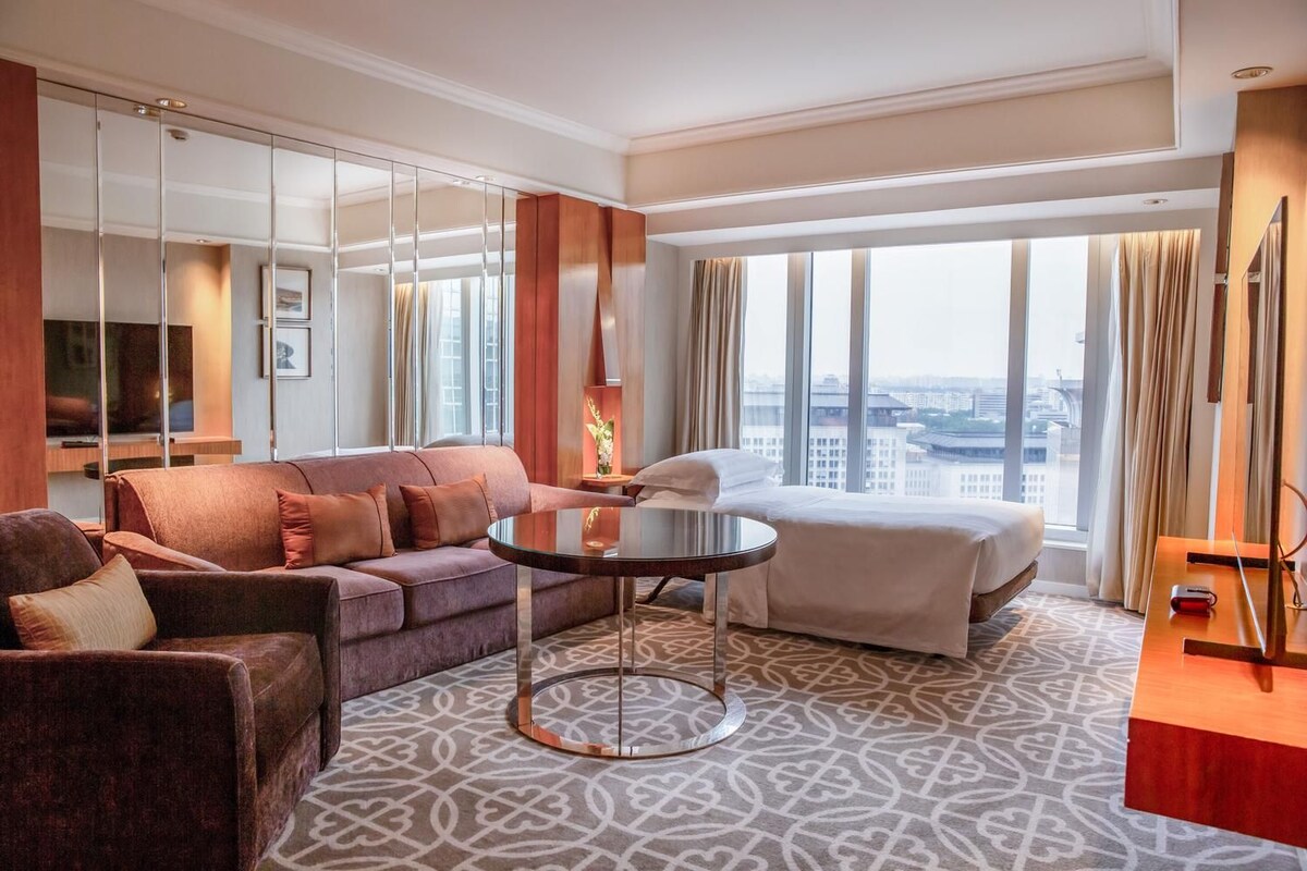 A hotel room with a sofa, a bed, a round table, and a large window offering a city view.