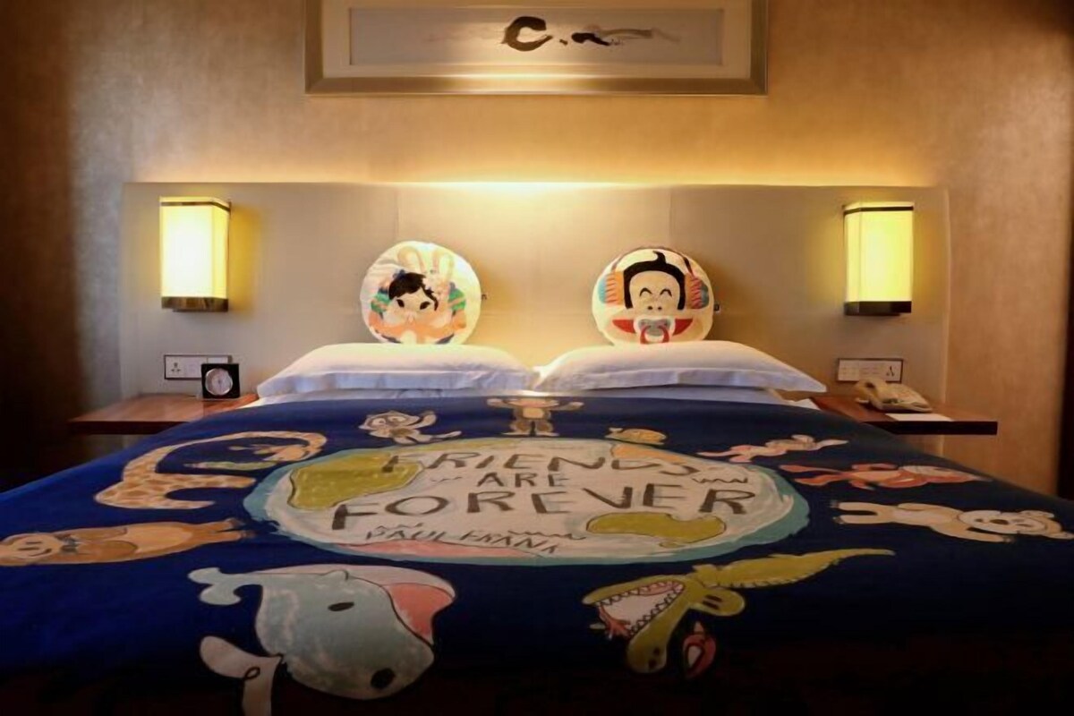 A hotel room with a bed featuring a 'Friends' themed bedspread, two wall-mounted lamps, and a framed artwork above the bed.