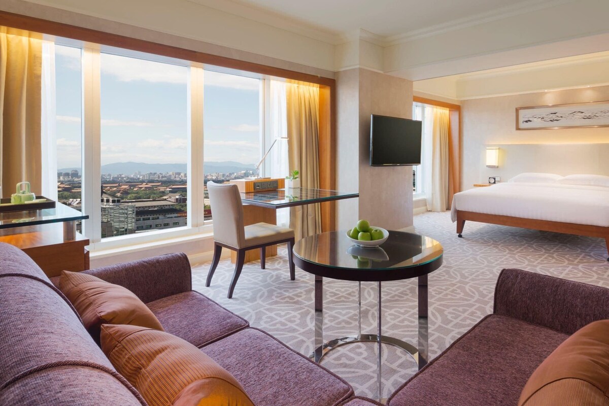 A hotel room with a large bed, a TV, a desk with a chair, and a sofa.