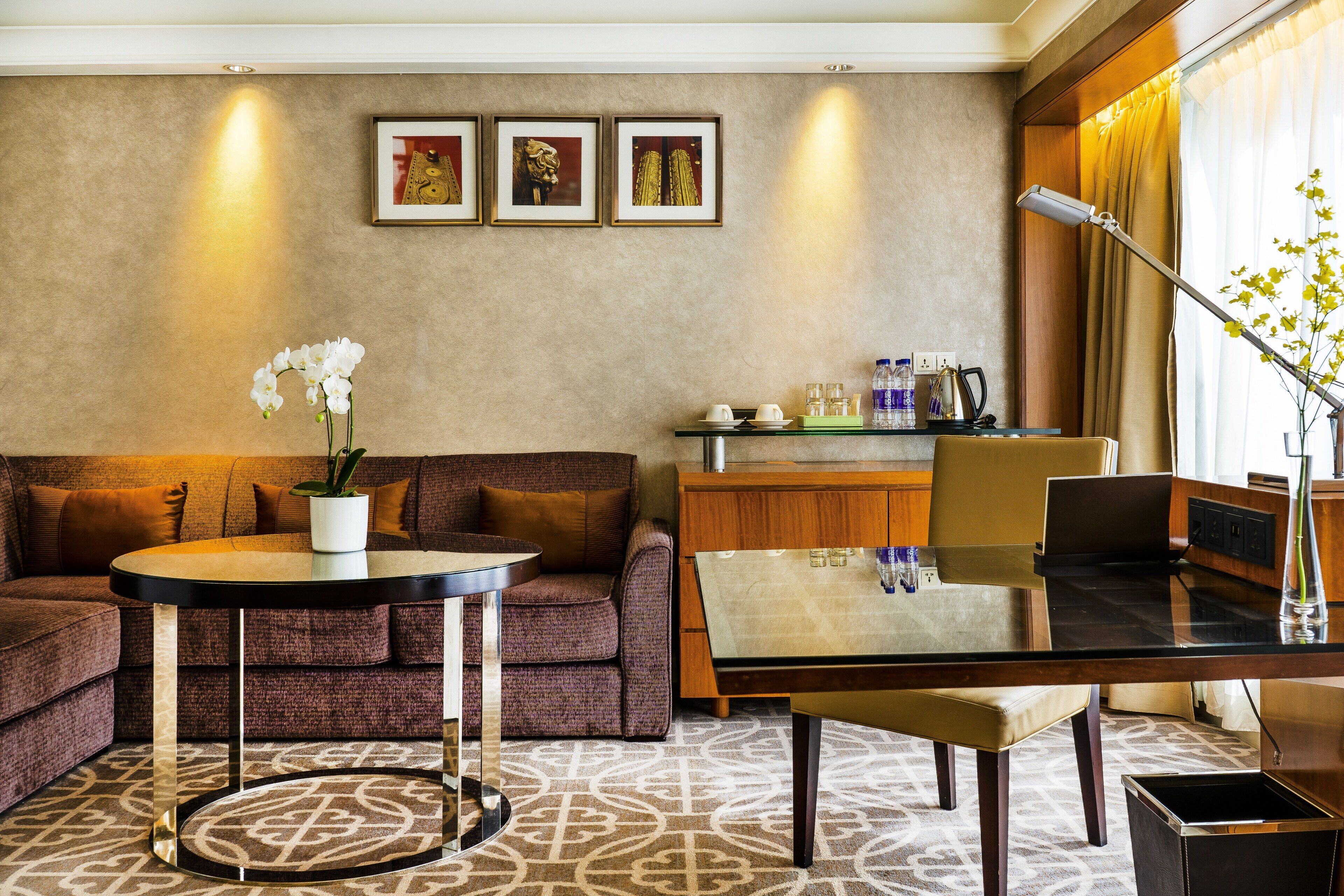 A modern hotel room with a sofa, a round table, a glass coffee table, a desk with a laptop, and a decorative vase.