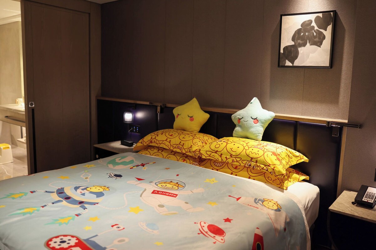 A hotel room with a bed featuring a colorful bedspread with cartoon characters, a bedside table with a lamp, and a wall-mounted artwork.