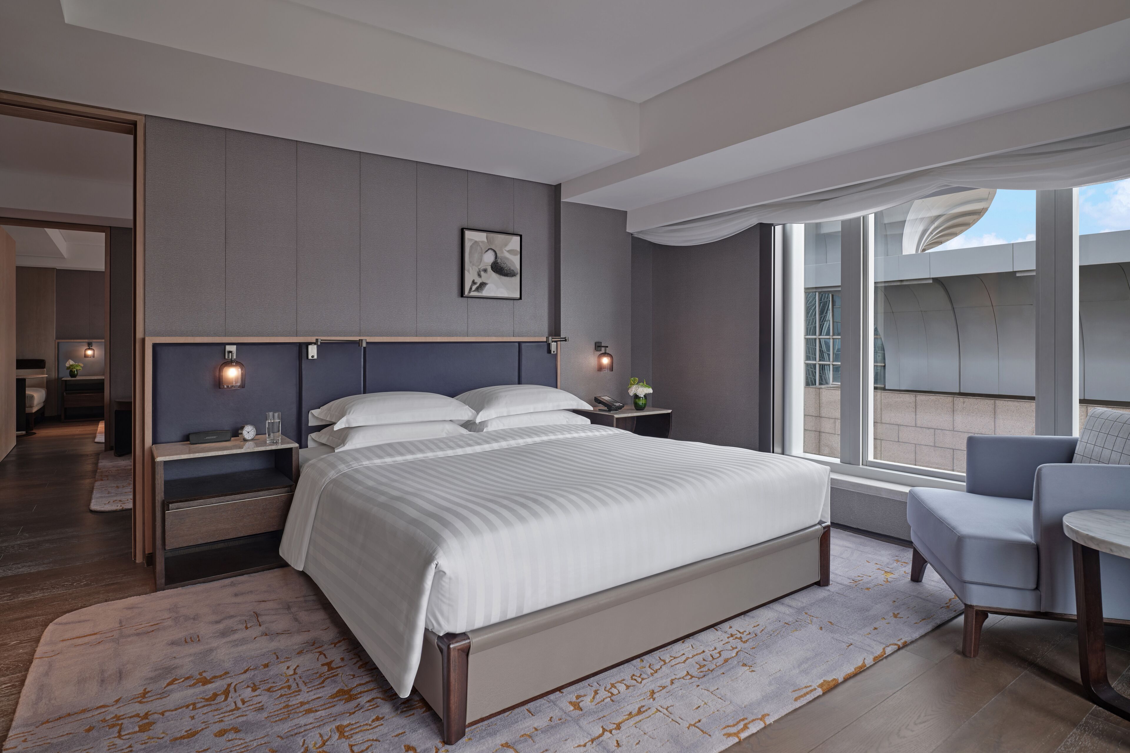 A modern bedroom with a large bed, bedside tables, a chair, and a view of the outside.