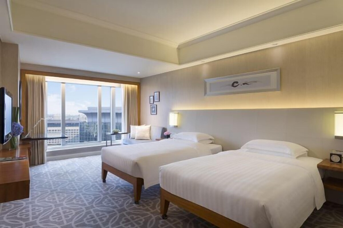 A hotel room with two beds, a large window with a city view, a desk with a lamp, and a framed picture on the wall.