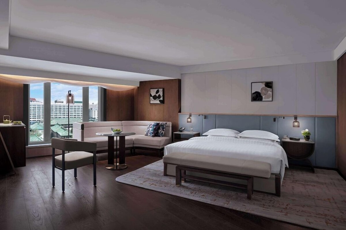 A modern hotel room with a large bed, a seating area with a sofa and chair, a small table, and a view of the city through large windows.