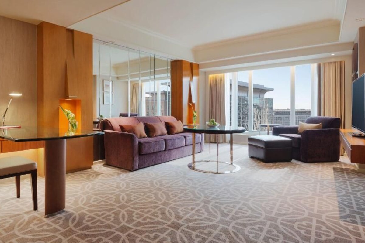 A modern hotel room with a glass dining table, a sofa, a chair, and a television.