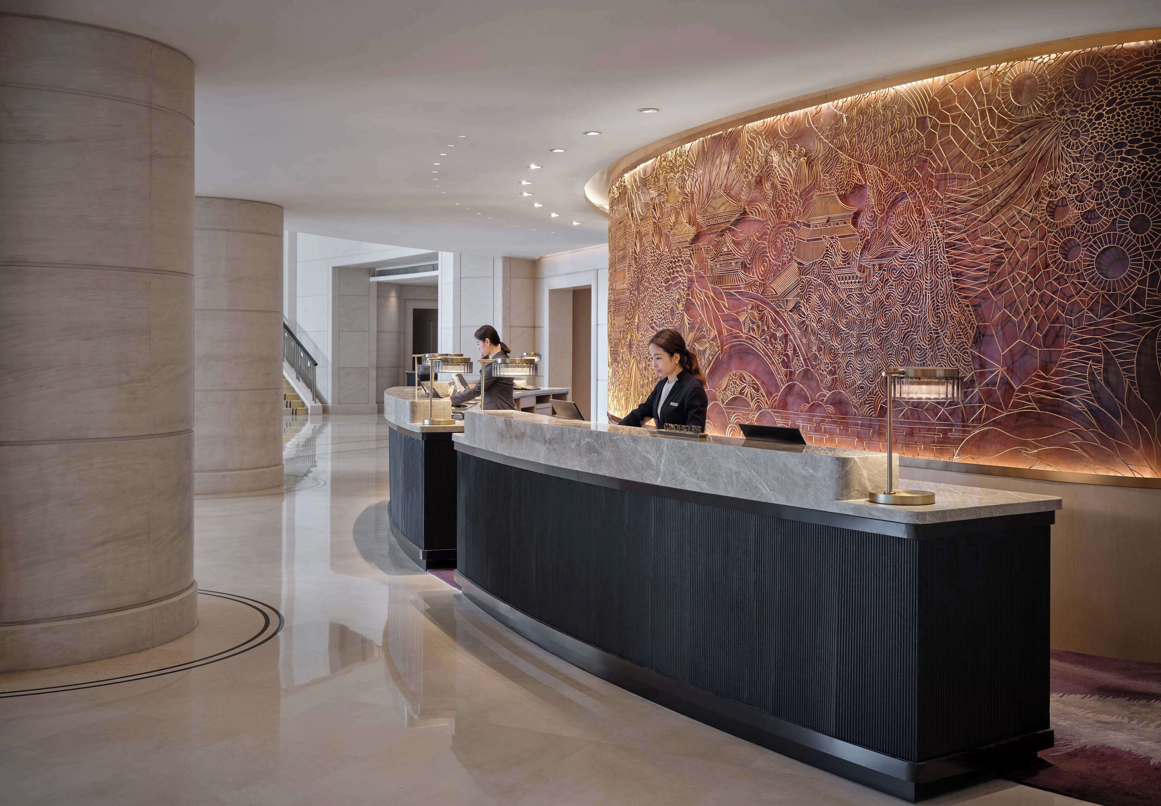 Zona de recepción moderna de un hotel, con un mostrador curvo, un gran elemento decorativo en la pared y una escalera al fondo.