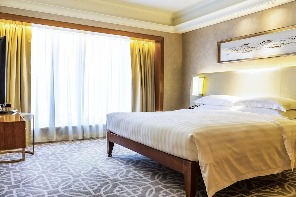 A hotel room with a bed, a desk, a lamp, and a large window with curtains.